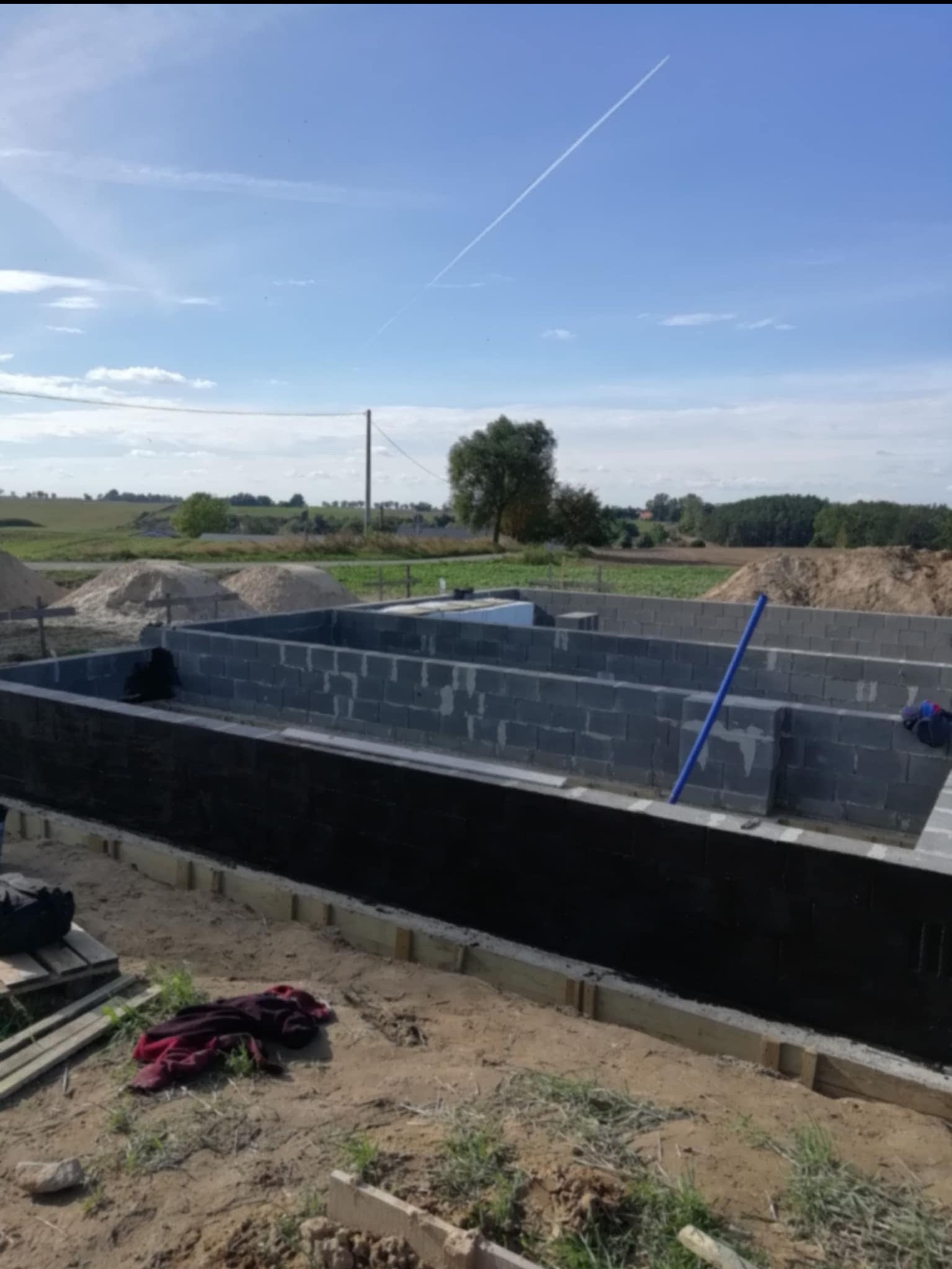 Fundamenty domu z bloczków betonowych, zabezpieczone hydroizolacją, szalunki drewniane, widok na okolicę z drzewami, błękitne niebo. Etap budowy.