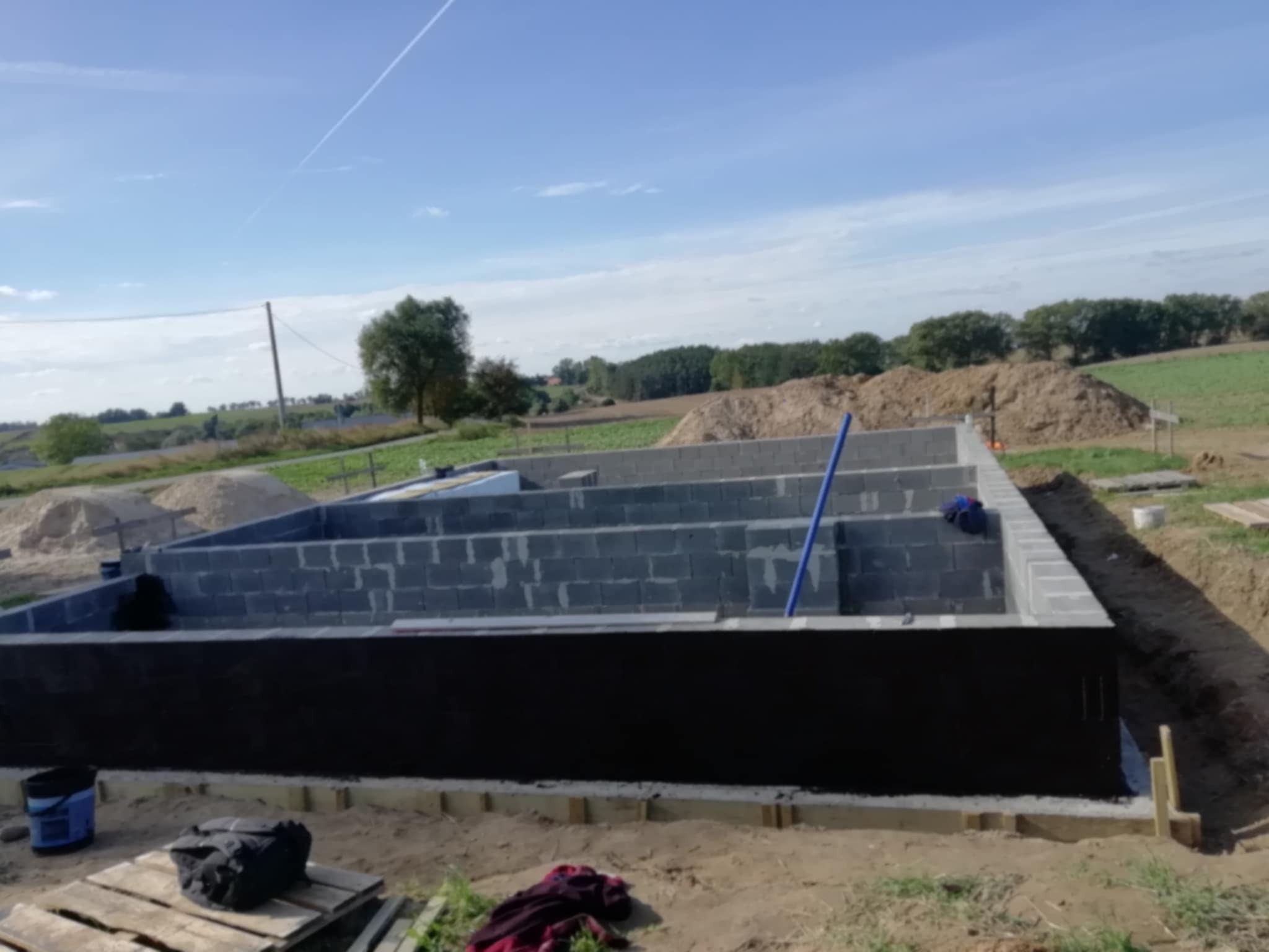 Wykopane fundamenty pod dom z izolacją przeciwwilgociową i częściowo wymurowanymi ścianami z bloczków betonowych na tle krajobrazu polnych wzgórz.