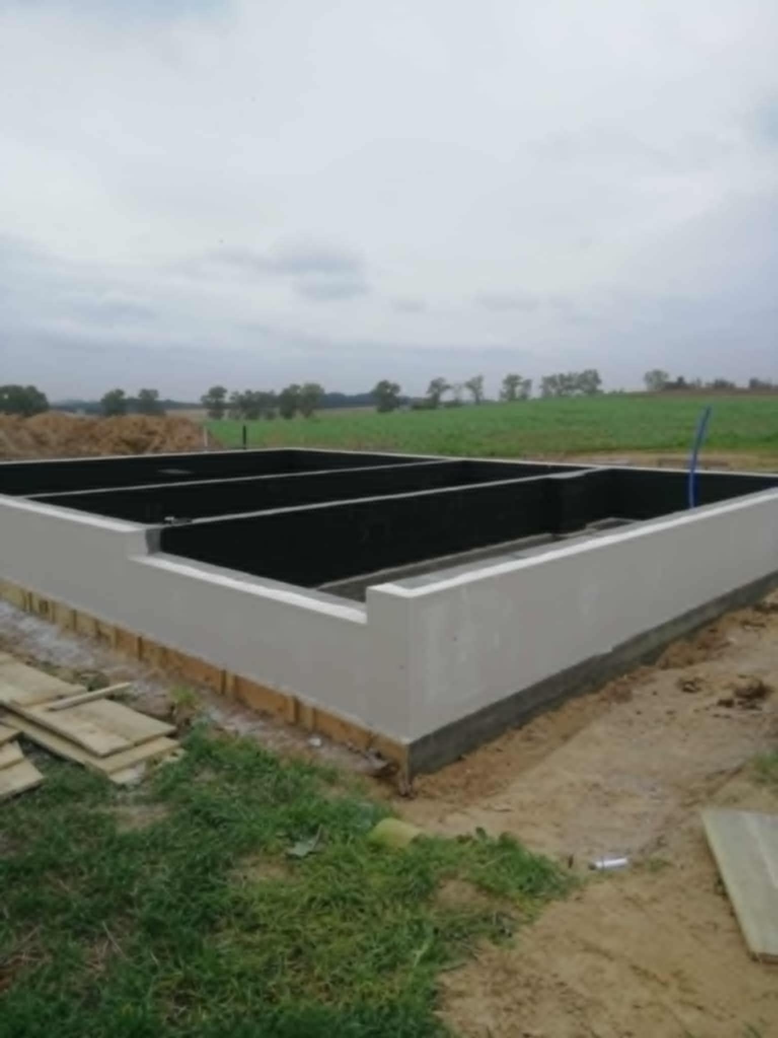 Zalane fundamenty domu z widoczną hydroizolacją wewnątrz, na zewnątrz drewniane deski i ziemia z trawą. W tle zielone pole i pochmurne niebo.