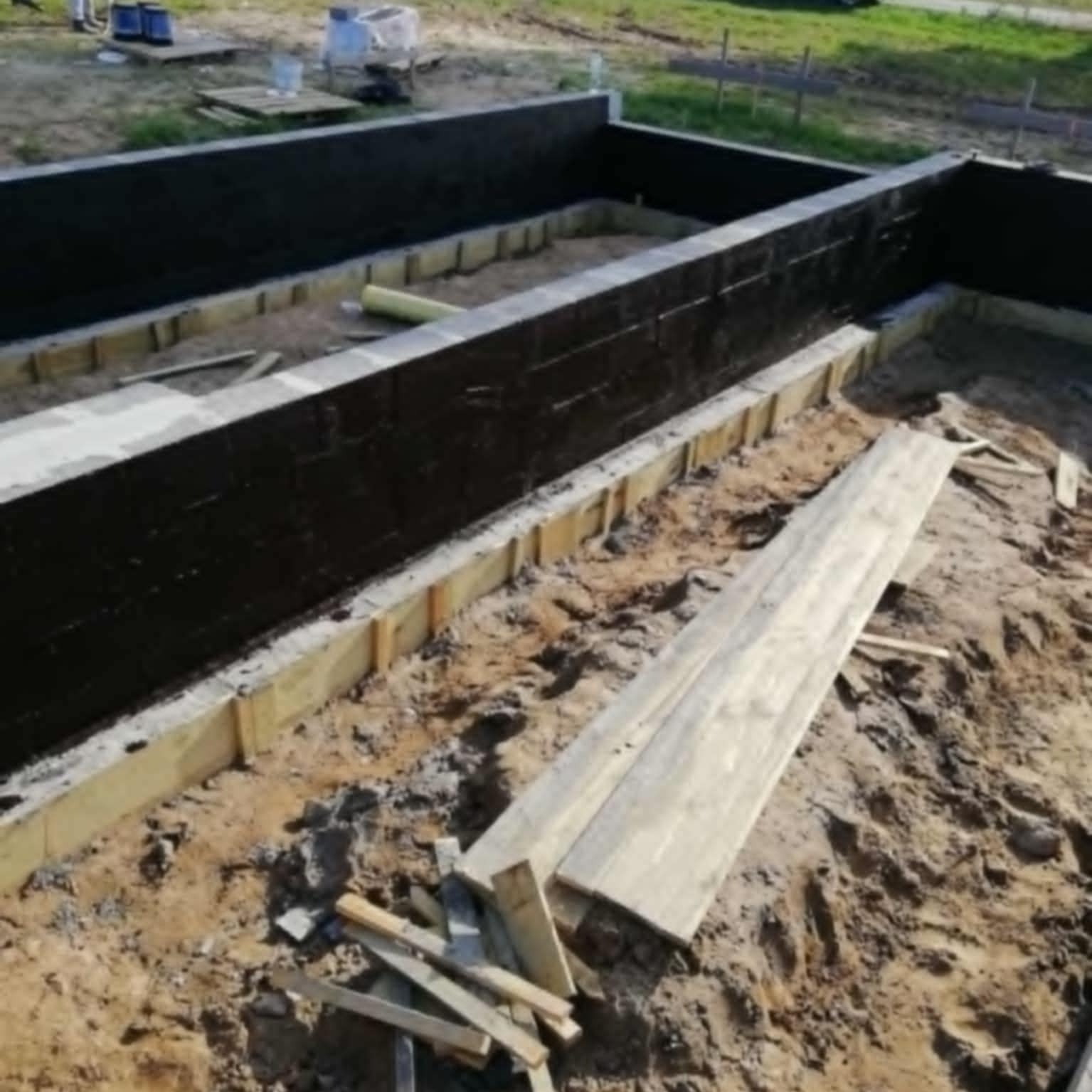 Zabezpieczone fundamenty domu w trakcie budowy, izolacja przeciwwilgociowa, szalunki drewniane, przygotowanie do dalszych prac. Widok z góry.