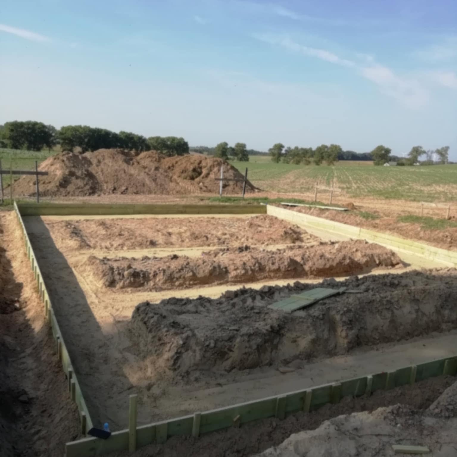 Fundamenty domu w trakcie budowy, widoczne drewniane szalunki na tle krajobrazu z polami i drzewami. Wykopane rowy pod ławy fundamentowe wypełnione ziemią.