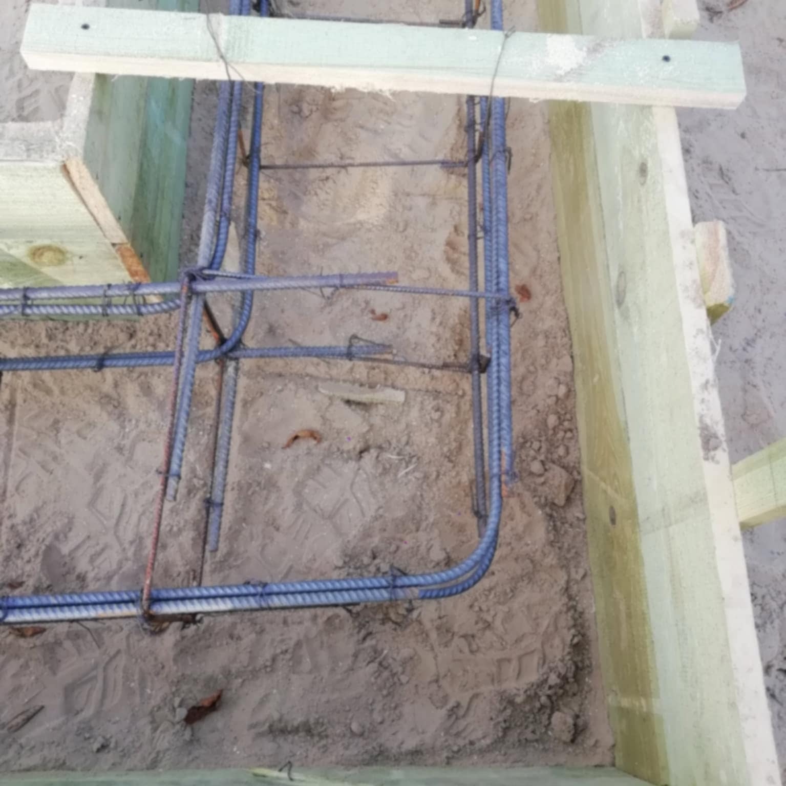 Zbrojenie fundamentów w drewnianym szalunku, widoczne pręty zbrojeniowe i ślady stóp w piasku. Przygotowanie do wylewania betonu.