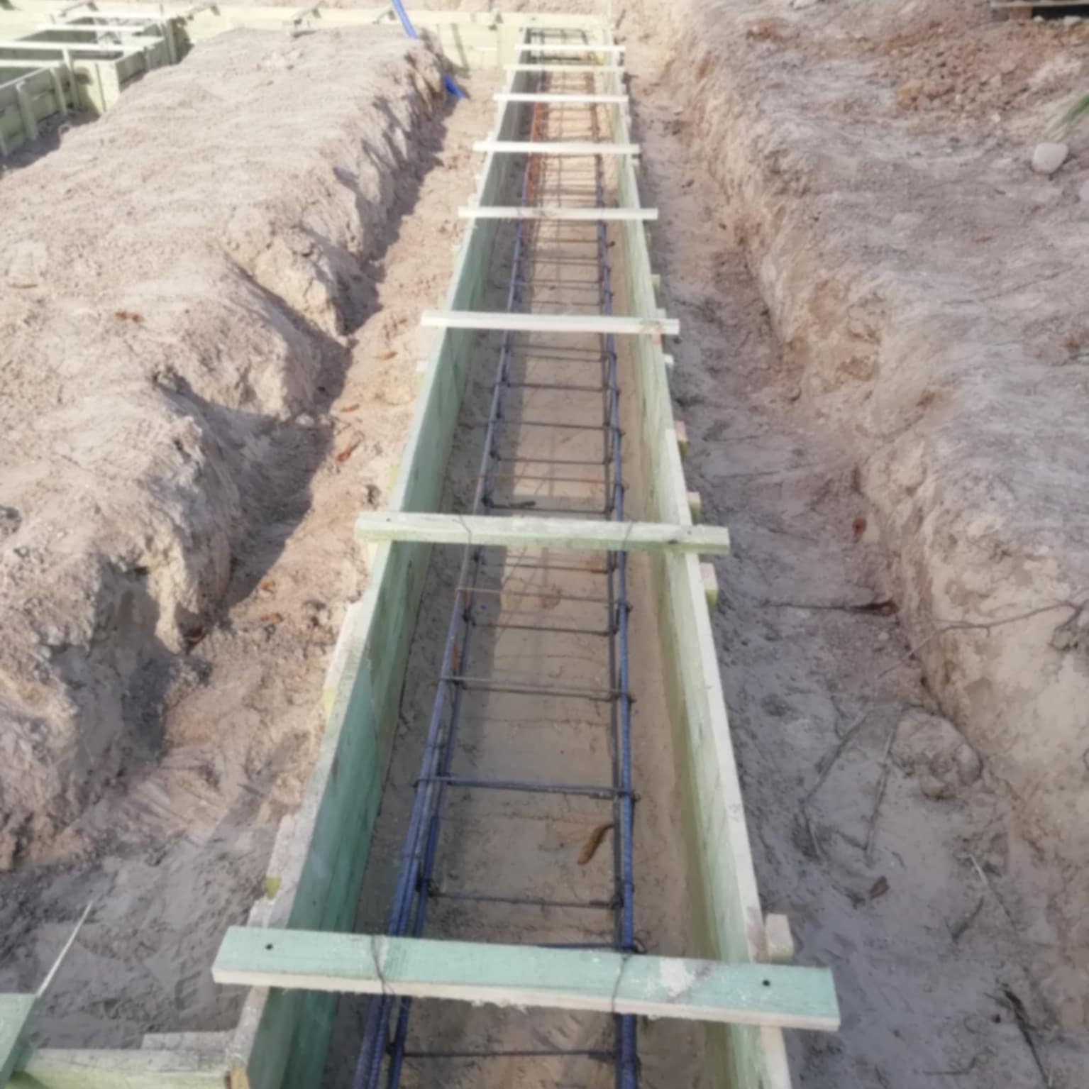 Przygotowanie szalunku i zbrojenia pod fundament. Widoczne drewniane deski i stalowe pręty zbrojeniowe w wykopie, gotowe do zalania betonem.