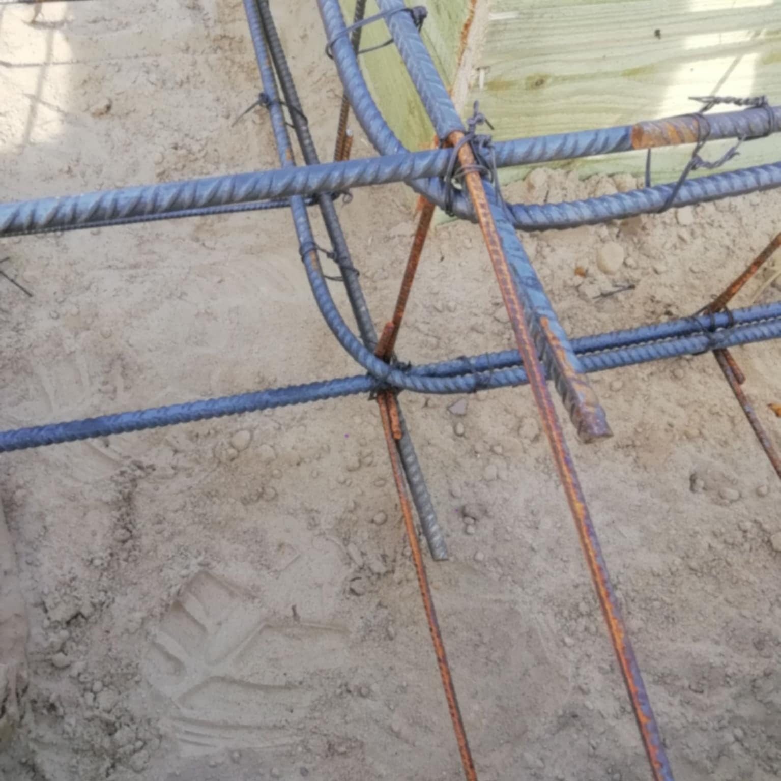 Zbrojenie fundamentu domu z bliska, widoczne pręty stalowe połączone drutem, w tle szalunek z desek i piasek. Detal z budowy domu.