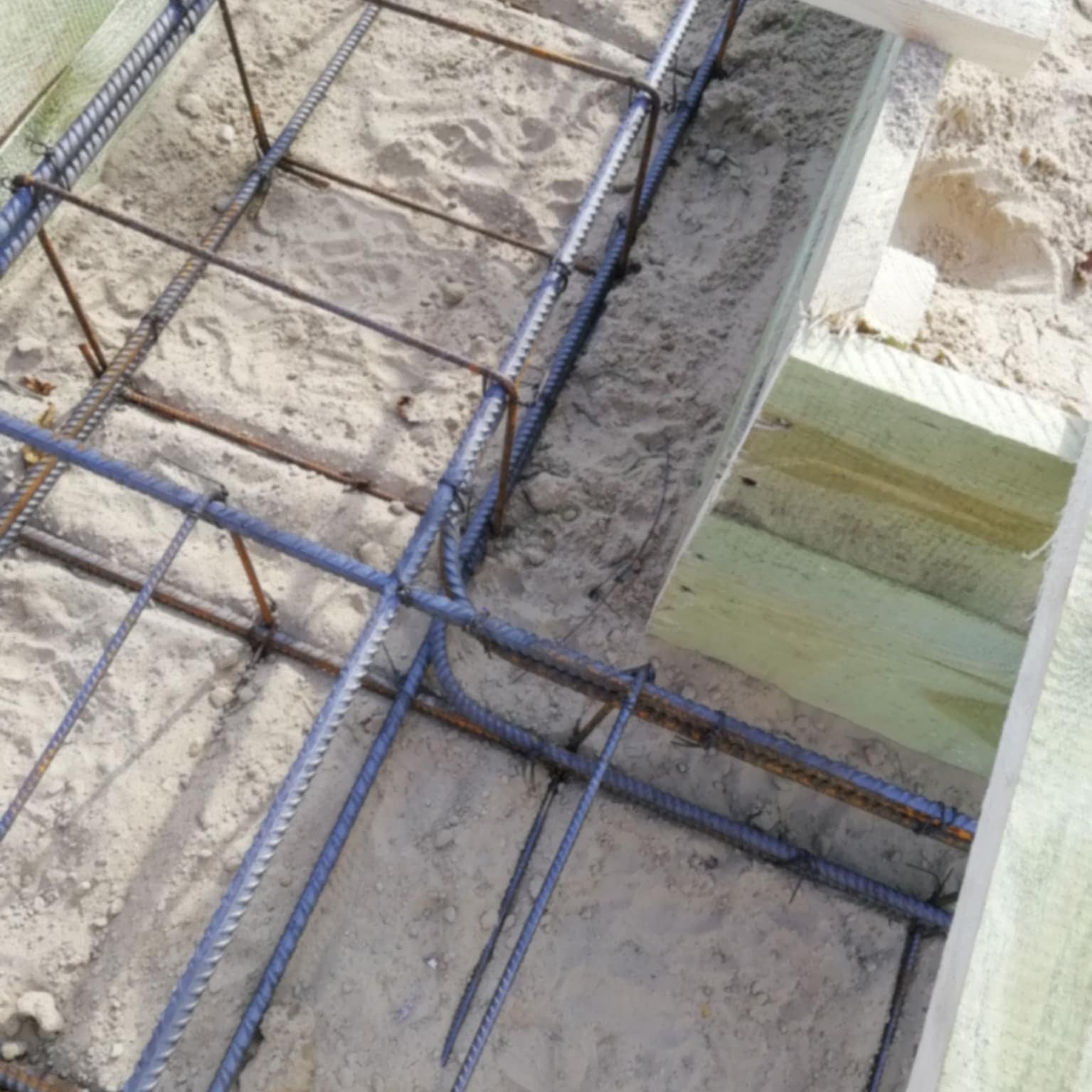 Zbrojenie fundamentów domu jednorodzinnego, widok z góry, szare pręty zbrojeniowe połączone drutem, drewniane szalunki w rogu kadru, piasek wewnątrz.