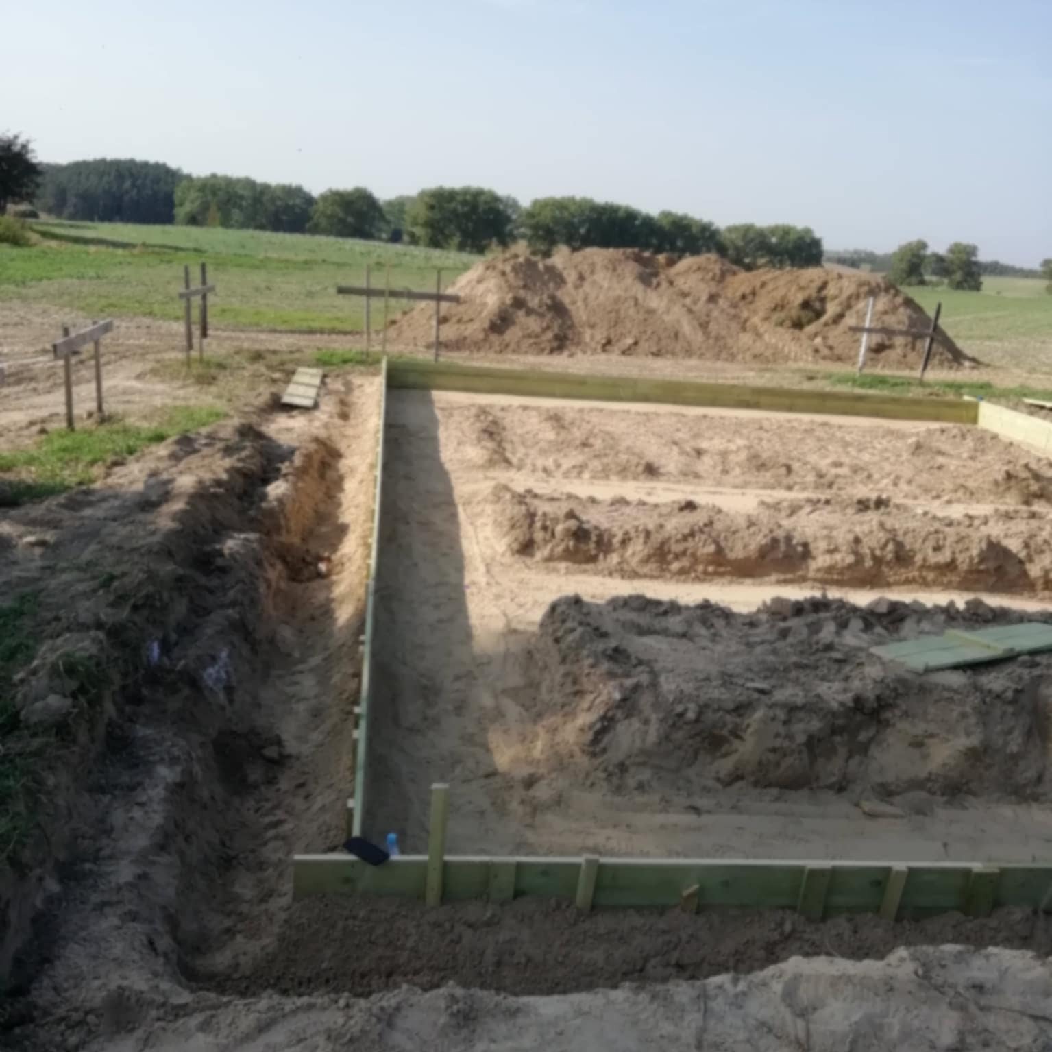 Fundamenty pod budowę domu w trakcie realizacji, widoczne drewniane szalunki i wykop, w tle krajobraz polny z drzewami.
