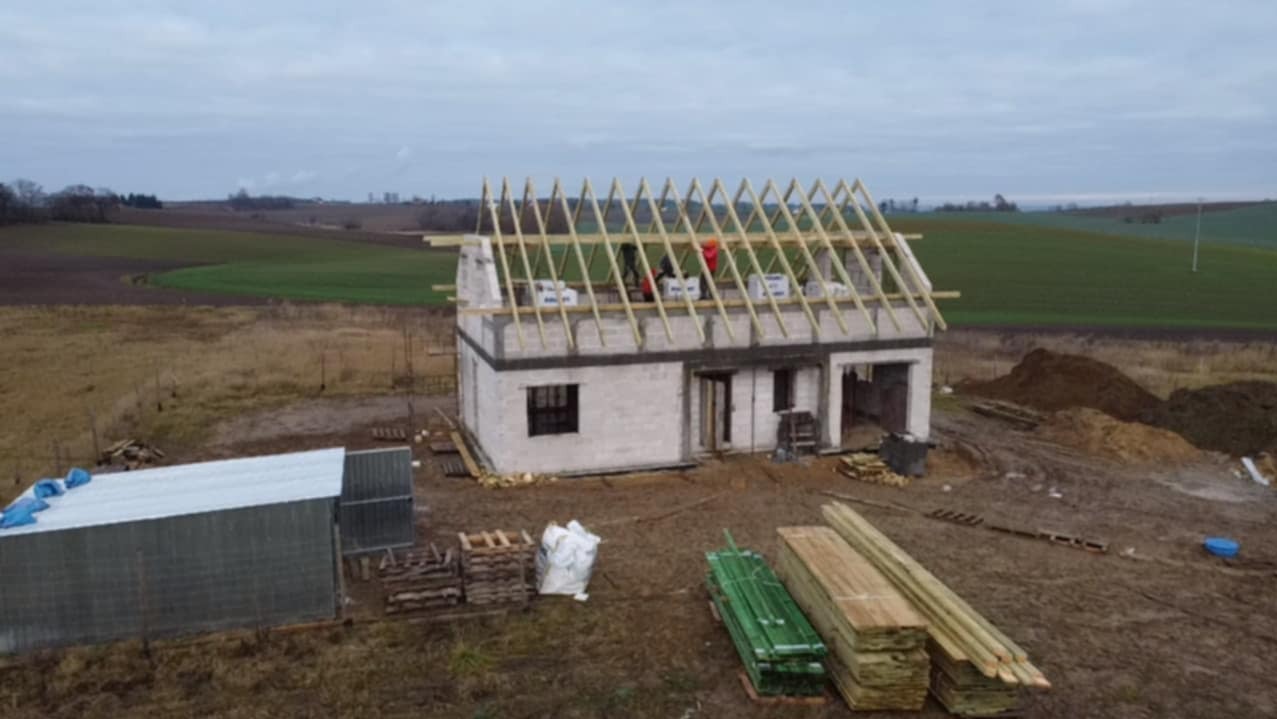 Budowa domu jednorodzinnego w stanie surowym otwartym: ściany z betonu komórkowego, drewniana konstrukcja dachu, materiały budowlane na działce. Widok z lotu ptaka.