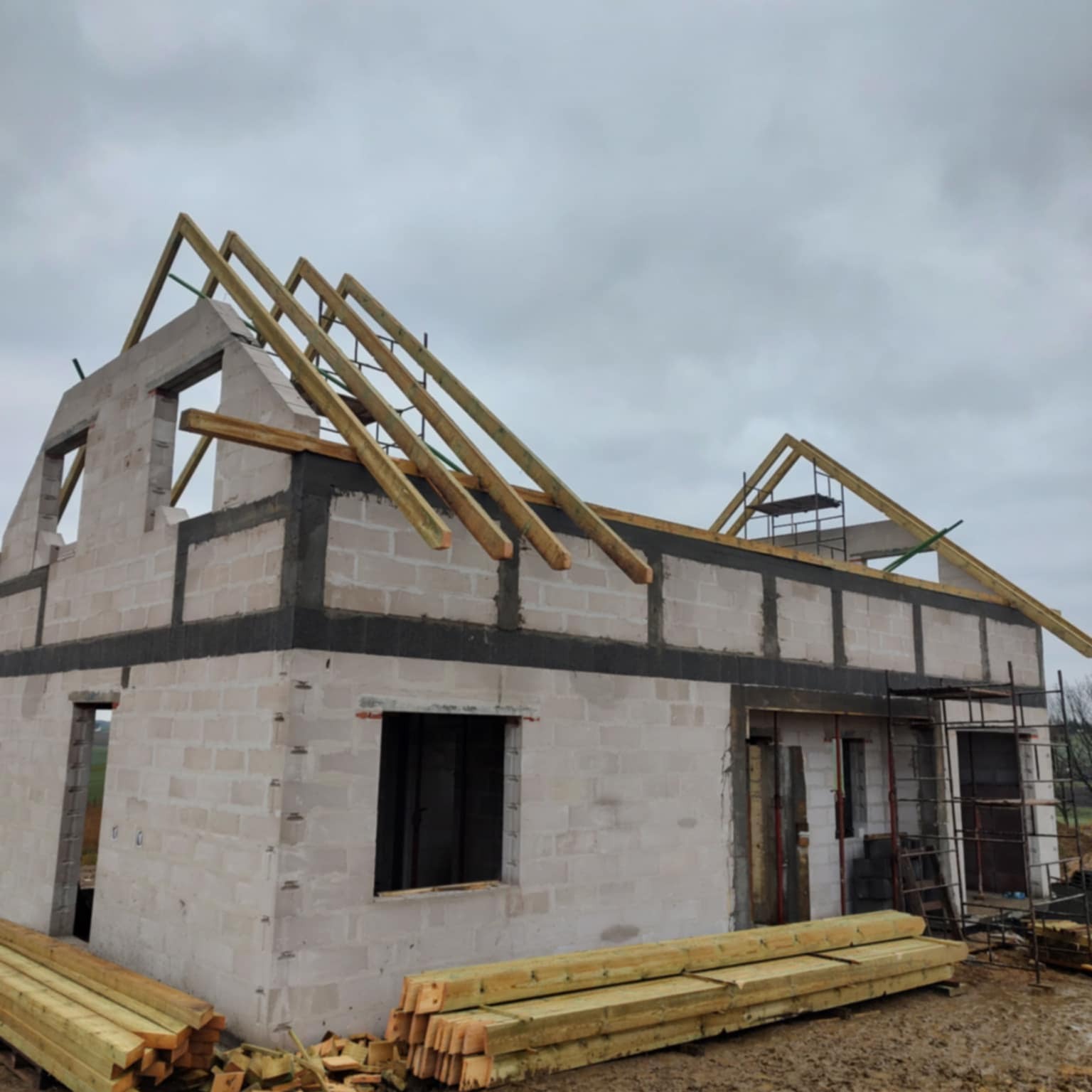 Budowa domu z bloczków: surowy stan, widoczne ściany, otwory okienne, drewniana konstrukcja dachu, rusztowania. Składowane drewno na pierwszym planie.