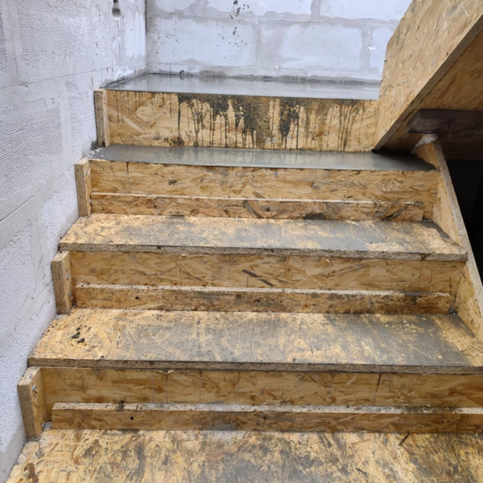 Surowe schody z płyty OSB w trakcie budowy. Widoczne betonowe wylewki na stopniach i surowe ściany z betonu komórkowego w tle. Detal budowlany.