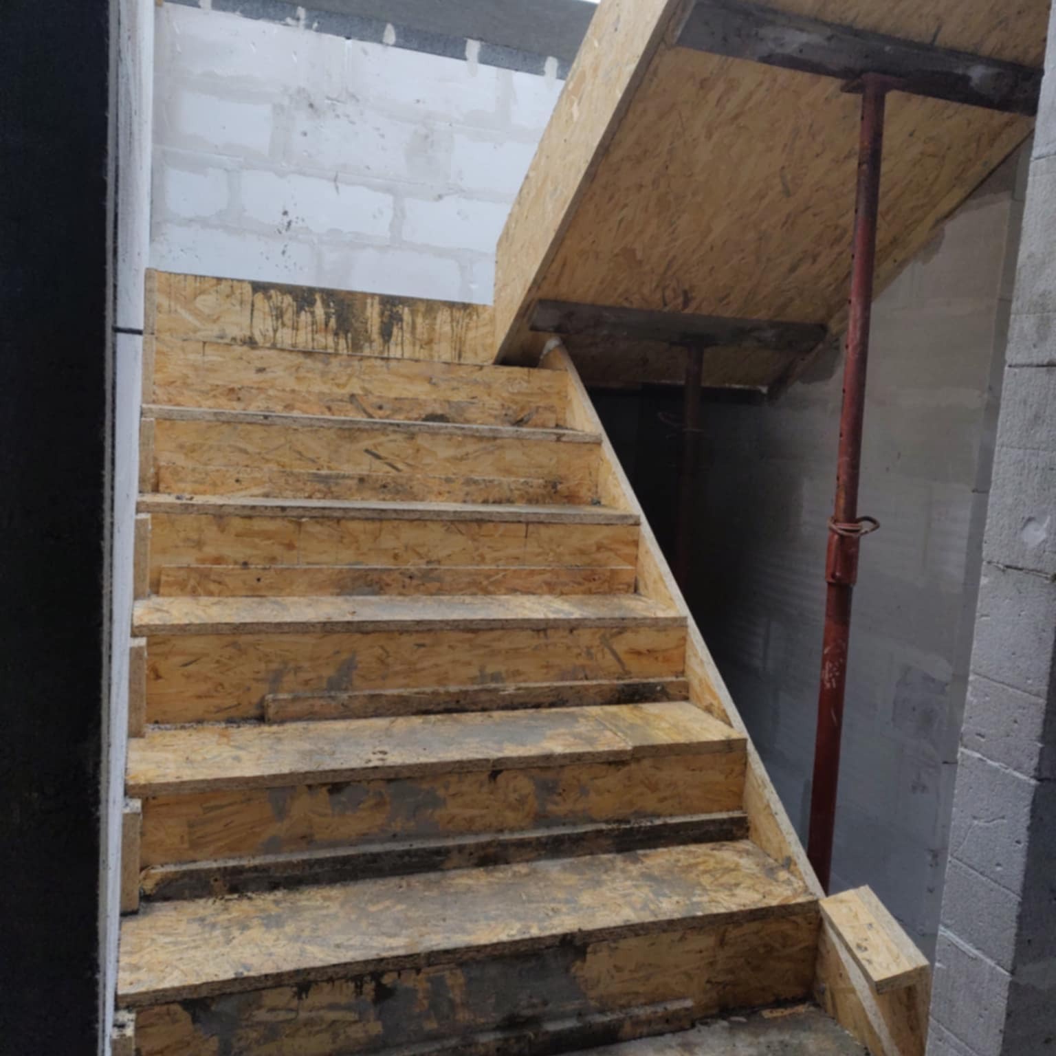 Tymczasowe, drewniane schody na budowie domu z bloczków, wsparte na metalowych stemplach. Widoczne ślady zaprawy i zabrudzenia.