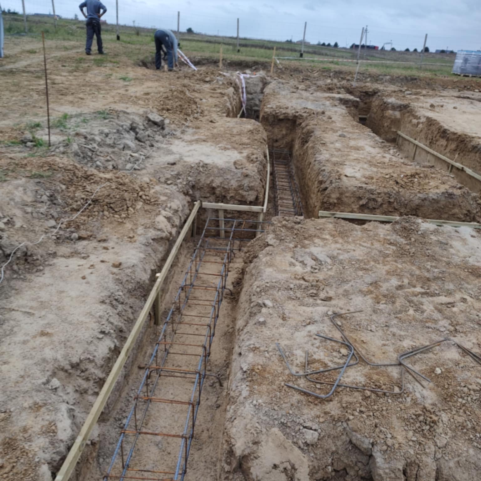 Zbrojenie fundamentów w wykopie, przygotowane do zalania betonem. Widoczne drewniane szalunki i pręty zbrojeniowe. W tle pracownicy budowlani i teren budowy.