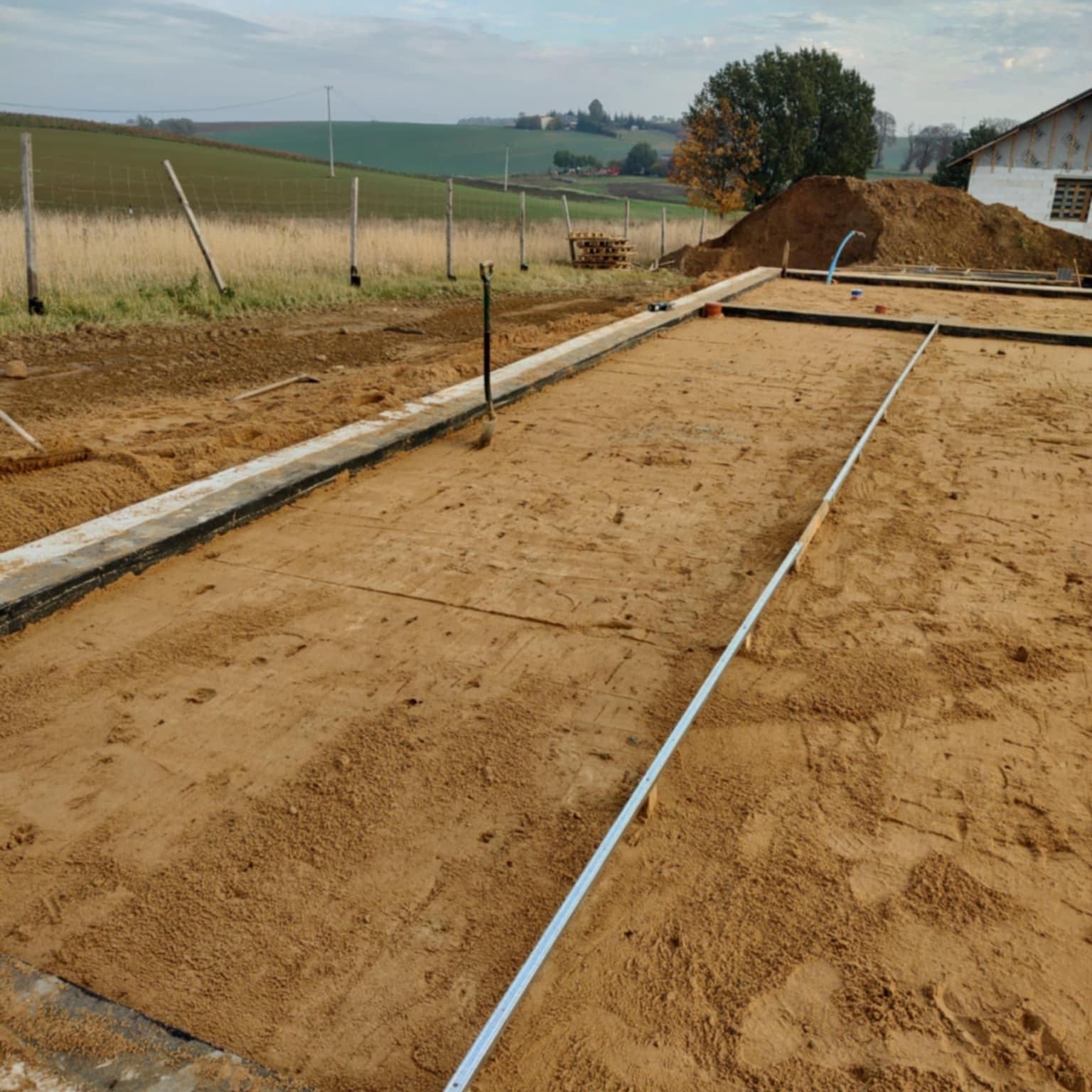 Przygotowany teren pod budowę domu z widocznymi fundamentami, ziemią i narzędziem. W tle krajobraz wiejski z drzewem i fragmentem budynku.