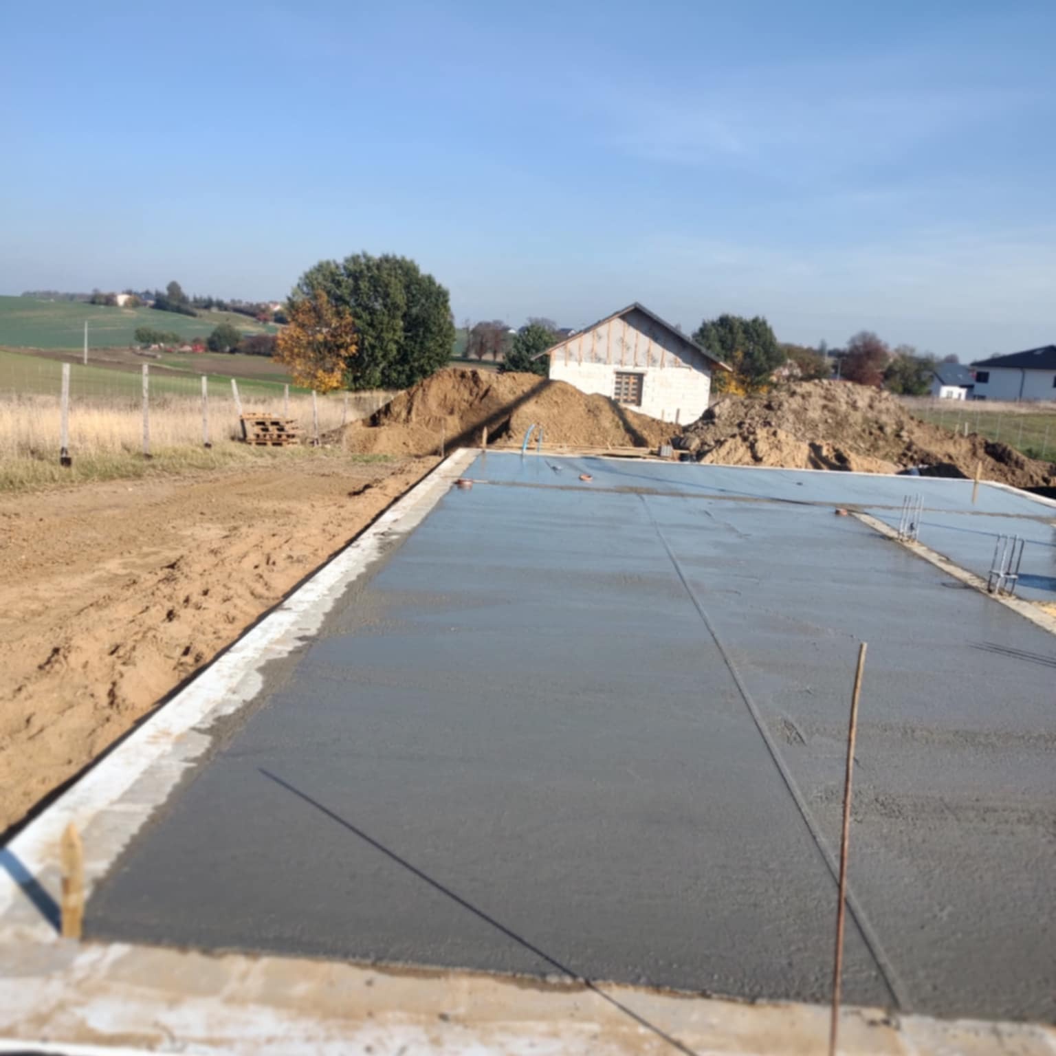 Wylana, świeża płyta fundamentowa pod budowę domu jednorodzinnego na tle krajobrazu wiejskiego w słoneczny dzień. Widoczne zbrojenia i szalunki.