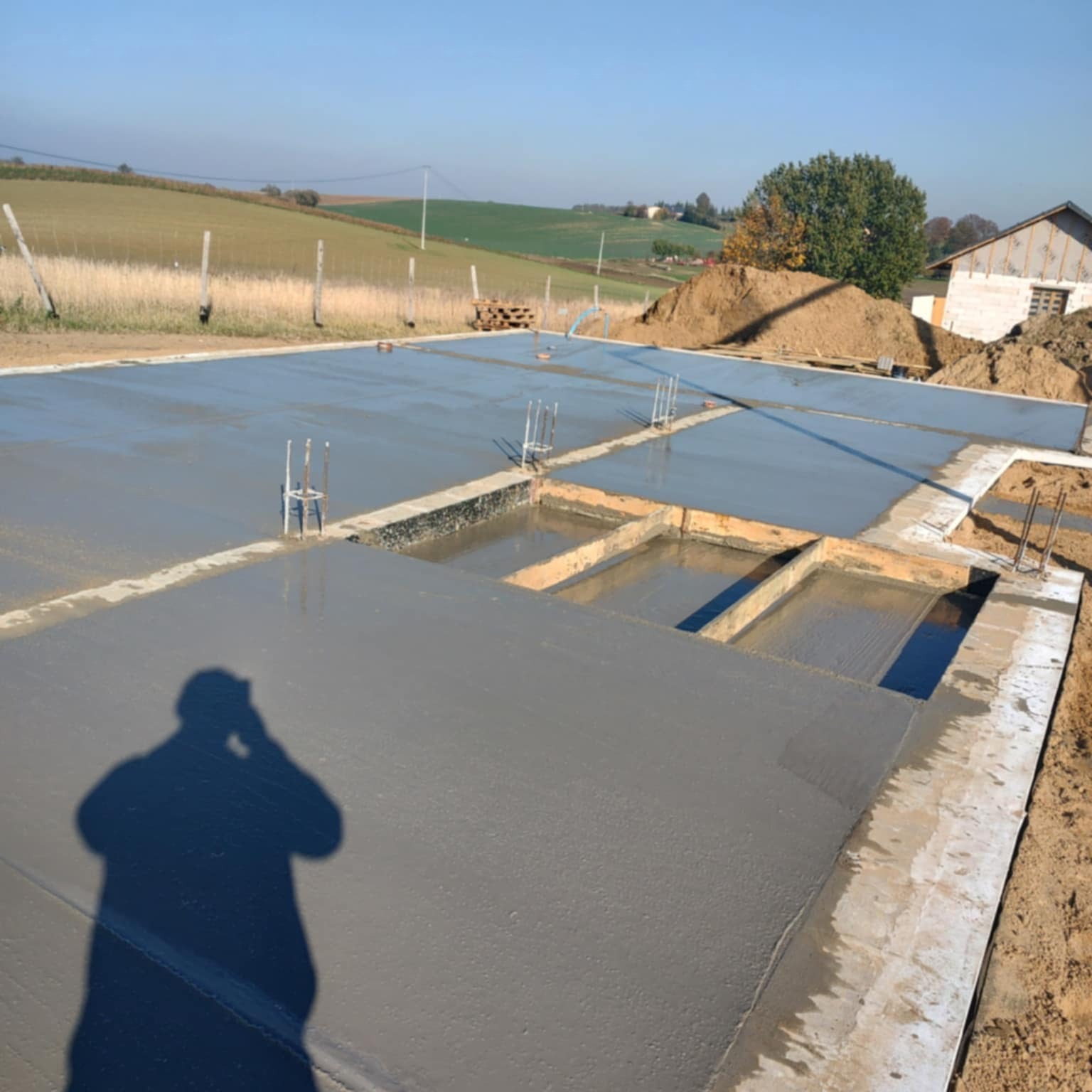 Świeżo wylana, gładka płyta fundamentowa z wystającymi prętami zbrojeniowymi, otoczona wykopem. W tle pagórkowaty krajobraz i częściowo widoczny budynek.