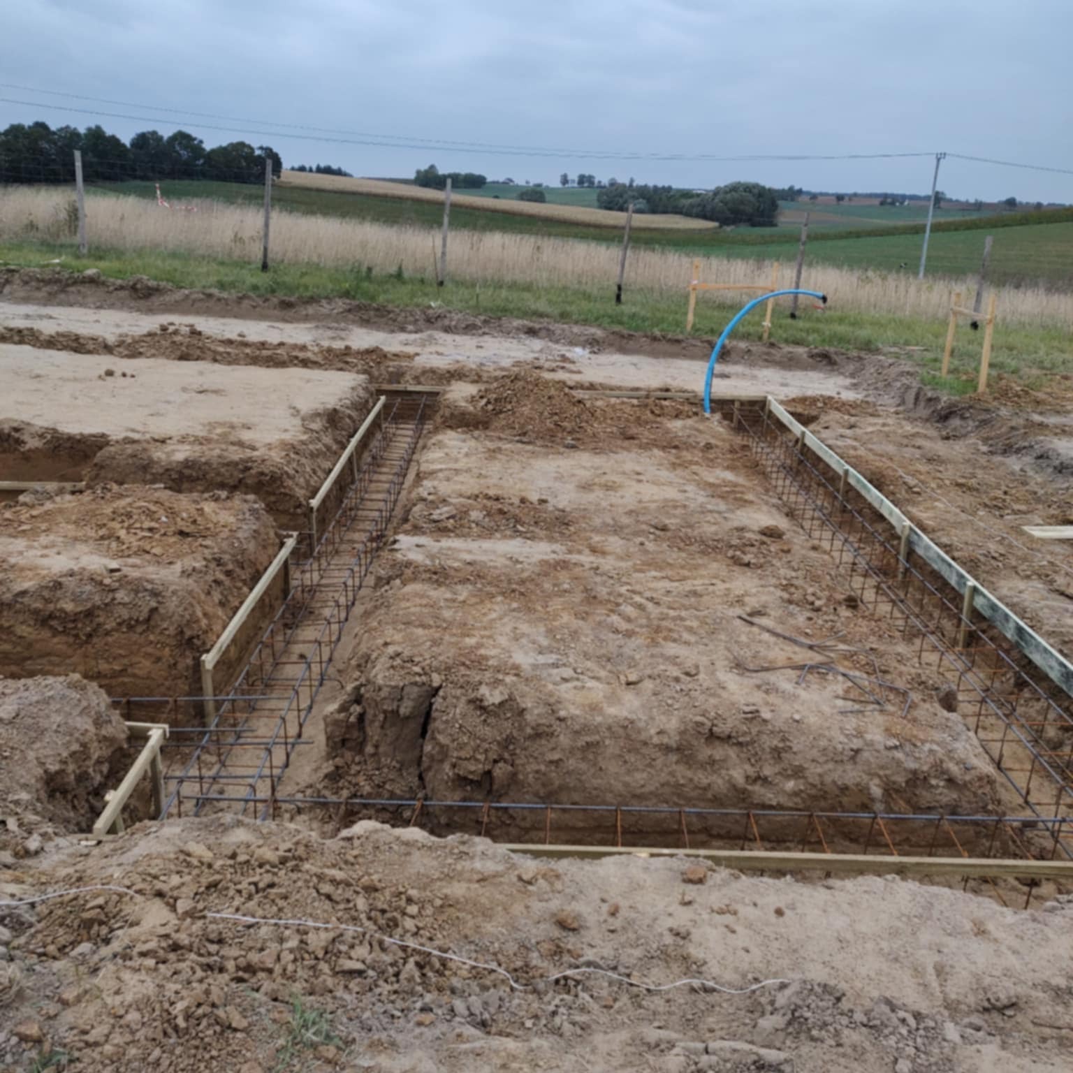 Przygotowanie fundamentów: wykopane rowy z drewnianym szalunkiem i zbrojeniem stalowym na tle krajobrazu polnych wzgórz pod pochmurnym niebem.