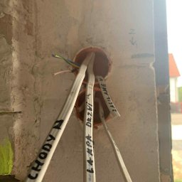 Eryk Kozak - Instalacja elektryczna w puszce podtynkowej, kable opisane markerem, surowa ściana z betonu komórkowego. Widoczne przewody: zielono-żółty, niebieski, brązowy.
