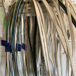 Eryk Kozak - Rozdzielnica elektryczna z poprowadzonymi przewodami o różnych kolorach, oznaczonymi niebieską taśmą, na tle surowej ściany z cegły.