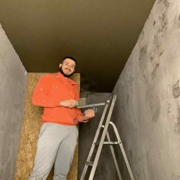 Eryk Kozak - Mężczyzna na drabinie z narzędziami do gładzi, po nałożeniu gładzi na sufit. Widoczne ściany w trakcie przygotowania do malowania.