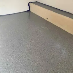 Szara, błyszcząca podłoga z żywicy epoksydowej z kolorowymi drobinkami, zabezpieczona taśmą malarską przy ścianie i cokole. Nowoczesne wykończenie podłogi.