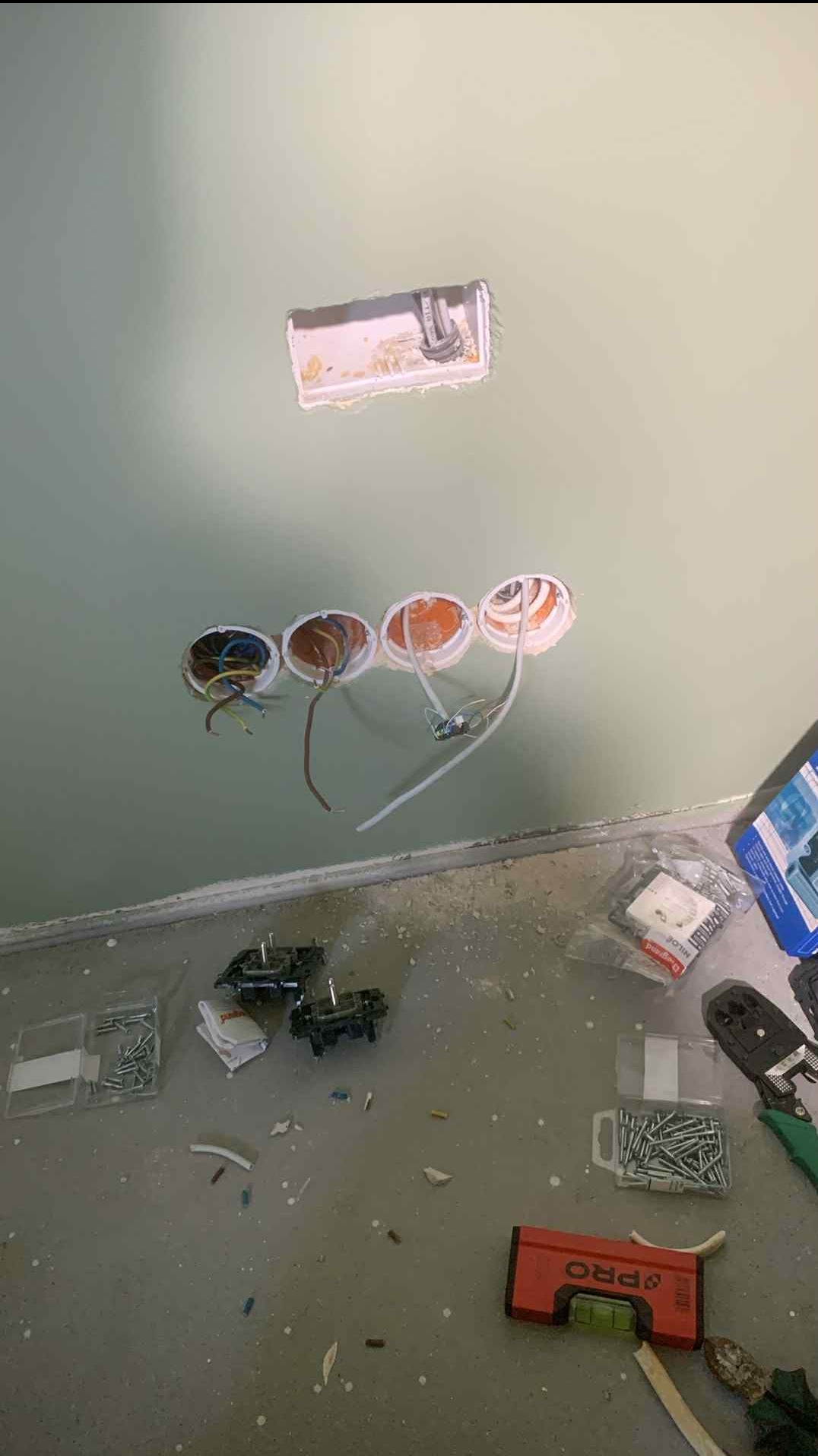 Ściana z wywierconymi otworami pod gniazdka elektryczne, widoczne kable, narzędzia i akcesoria elektryczne na podłodze, w trakcie prac instalacyjnych.