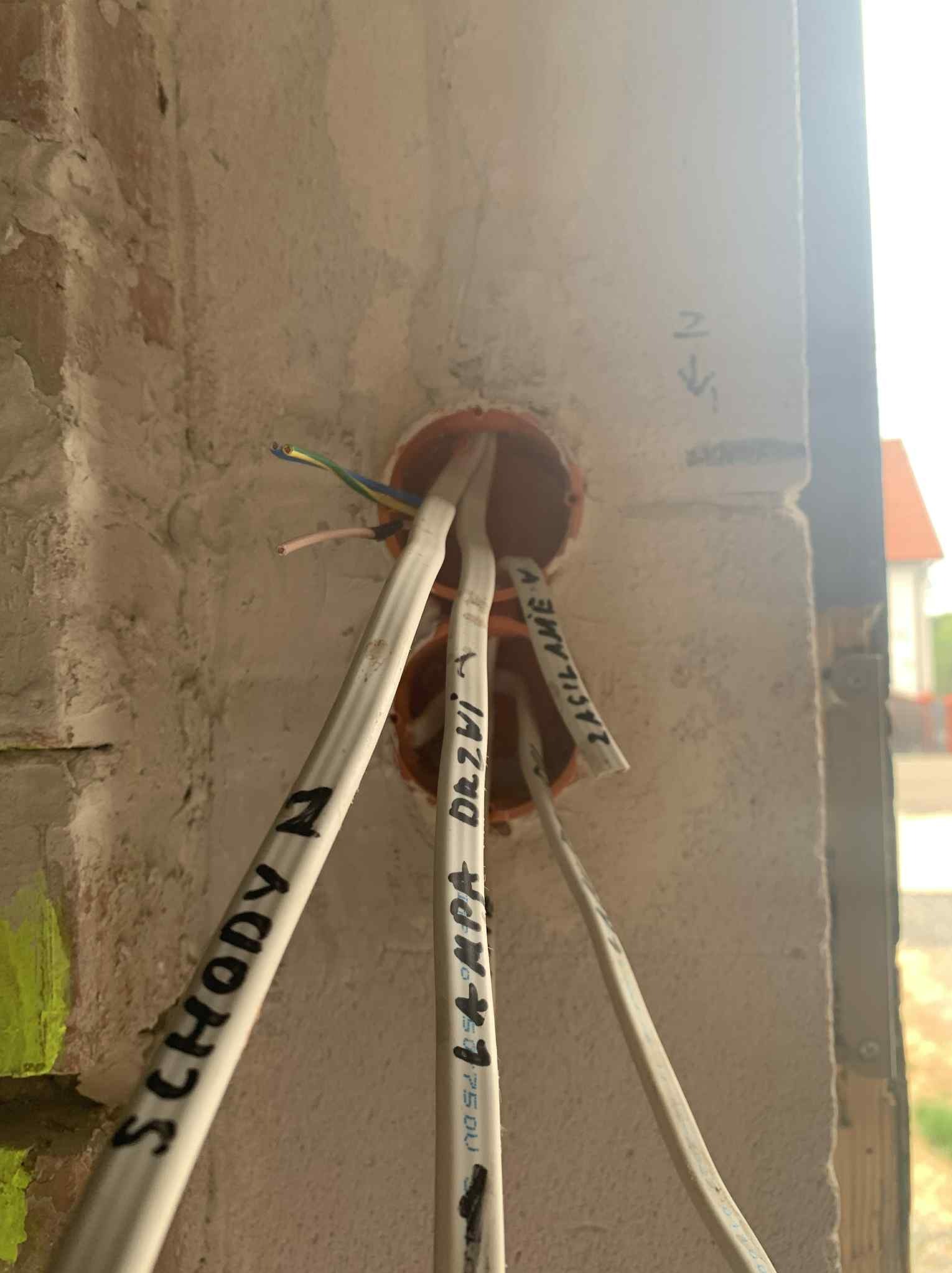 Instalacja elektryczna w puszce podtynkowej, kable opisane markerem, surowa ściana z betonu komórkowego. Widoczne przewody: zielono-żółty, niebieski, brązowy.