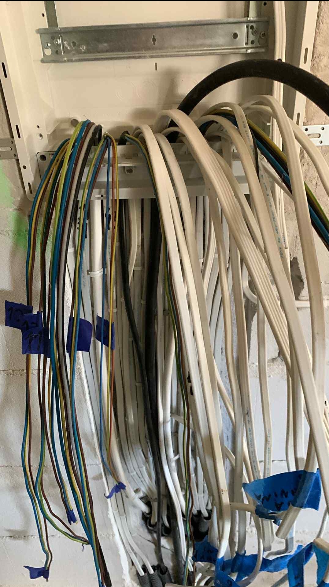 Rozdzielnica elektryczna z poprowadzonymi przewodami o różnych kolorach, oznaczonymi niebieską taśmą, na tle surowej ściany z cegły.