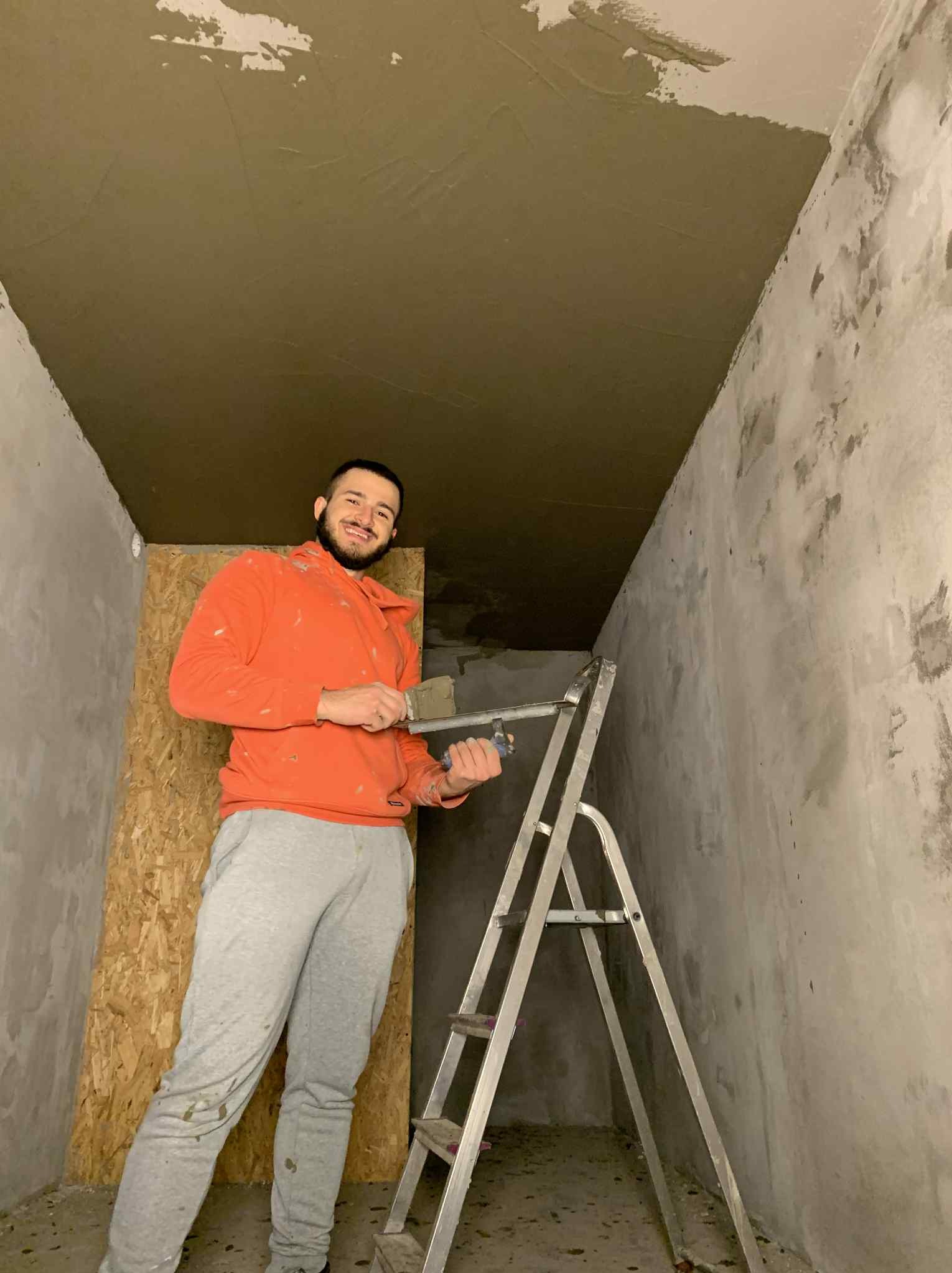 Mężczyzna na drabinie z narzędziami do gładzi, po nałożeniu gładzi na sufit. Widoczne ściany w trakcie przygotowania do malowania.