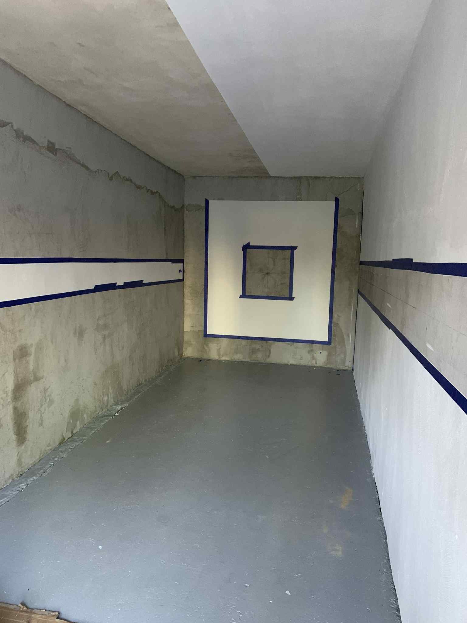 Przygotowane do malowania pomieszczenie z wyklejonymi taśmą fragmentami ścian i podłogi. Widoczne surowe i pomalowane fragmenty. Surowy beton na ścianach.