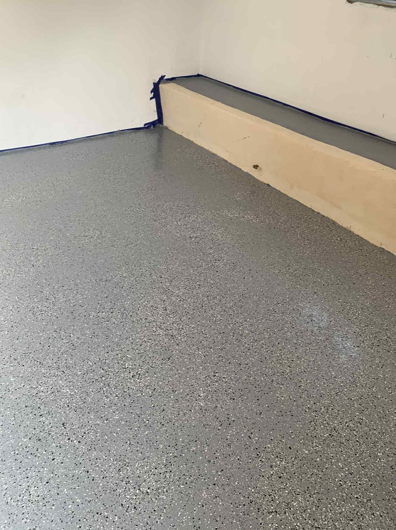 Szara, błyszcząca podłoga z żywicy epoksydowej z kolorowymi drobinkami, zabezpieczona taśmą malarską przy ścianie i cokole. Nowoczesne wykończenie podłogi.