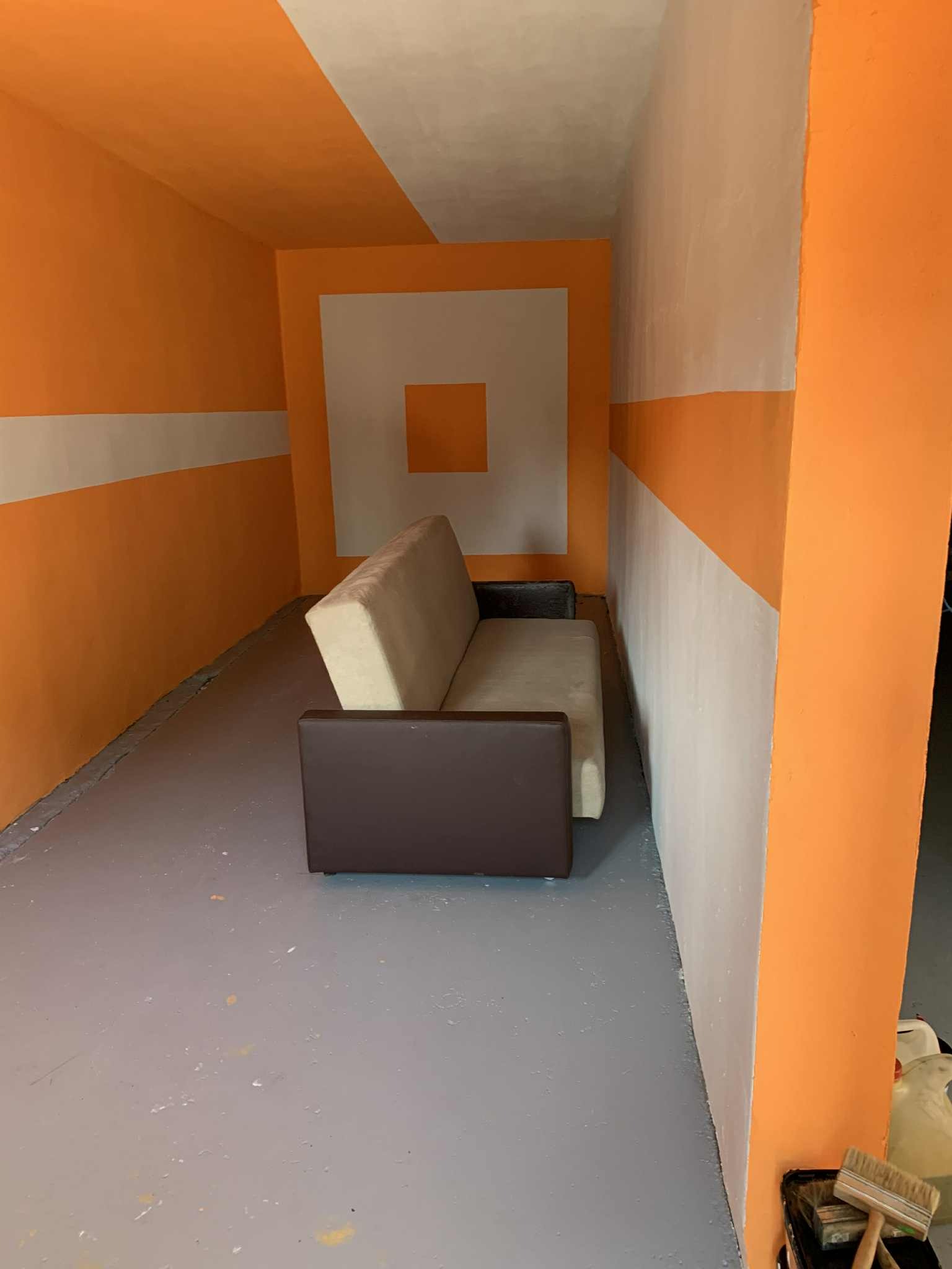 Wnętrze po malowaniu: pomarańczowo-białe ściany z geometrycznymi wzorami, szara podłoga z plamami farby i sofa. Widoczne pędzle malarskie.