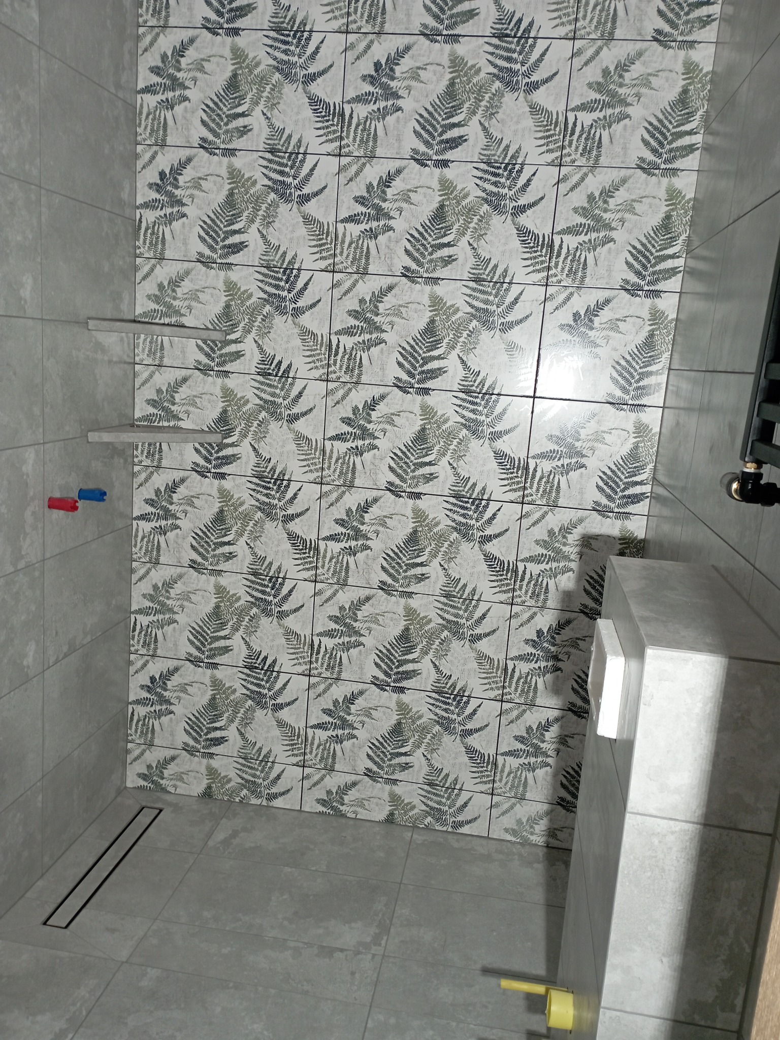 Wykończona łazienka z szarymi płytkami i motywem roślinnym na ścianie. Widoczny odpływ liniowy, półki i fragment toalety podtynkowej.