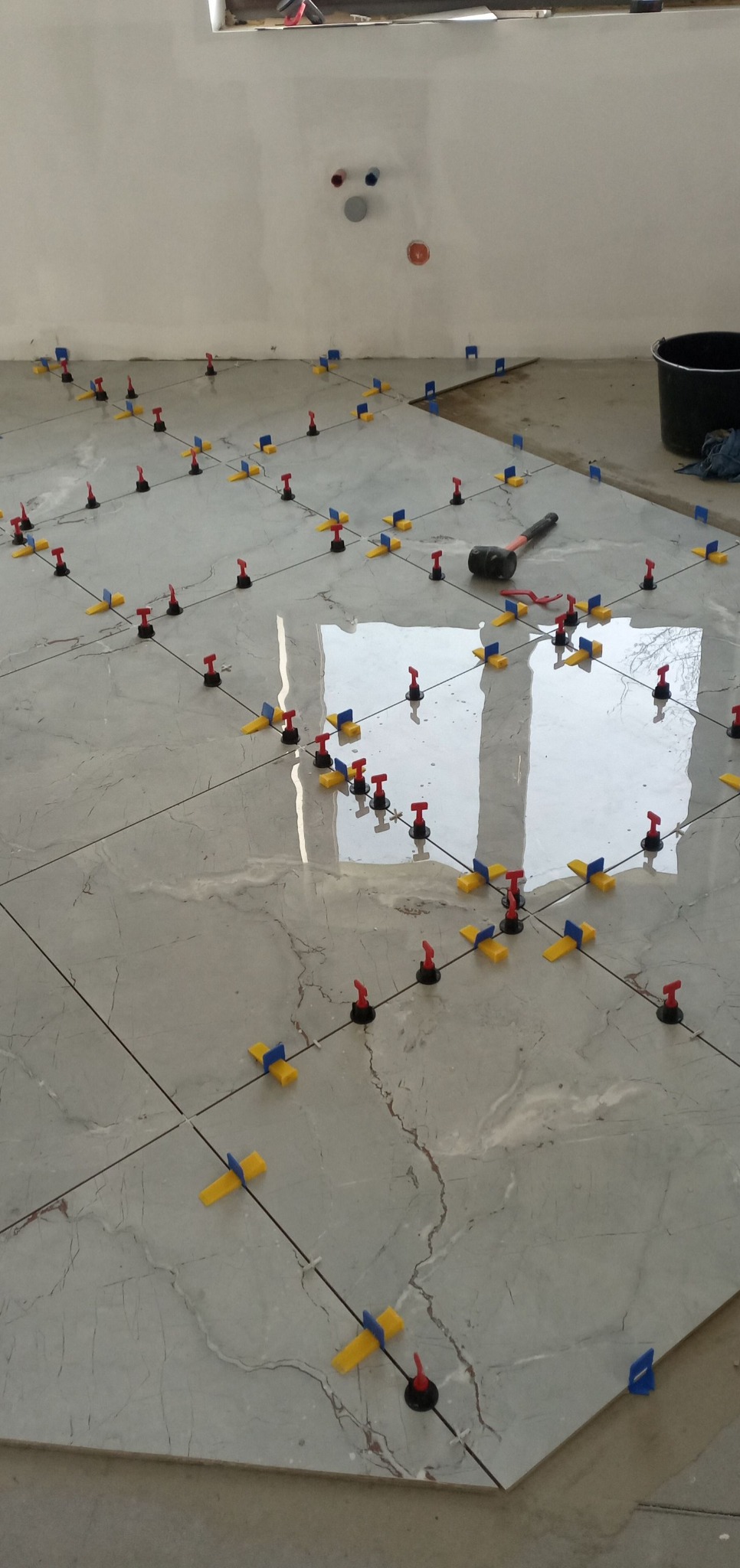 Położone płytki podłogowe z systemem poziomowania, widoczny młotek gumowy i wiadro. Szare płytki imitujące marmur, oświetlone naturalnym światłem.