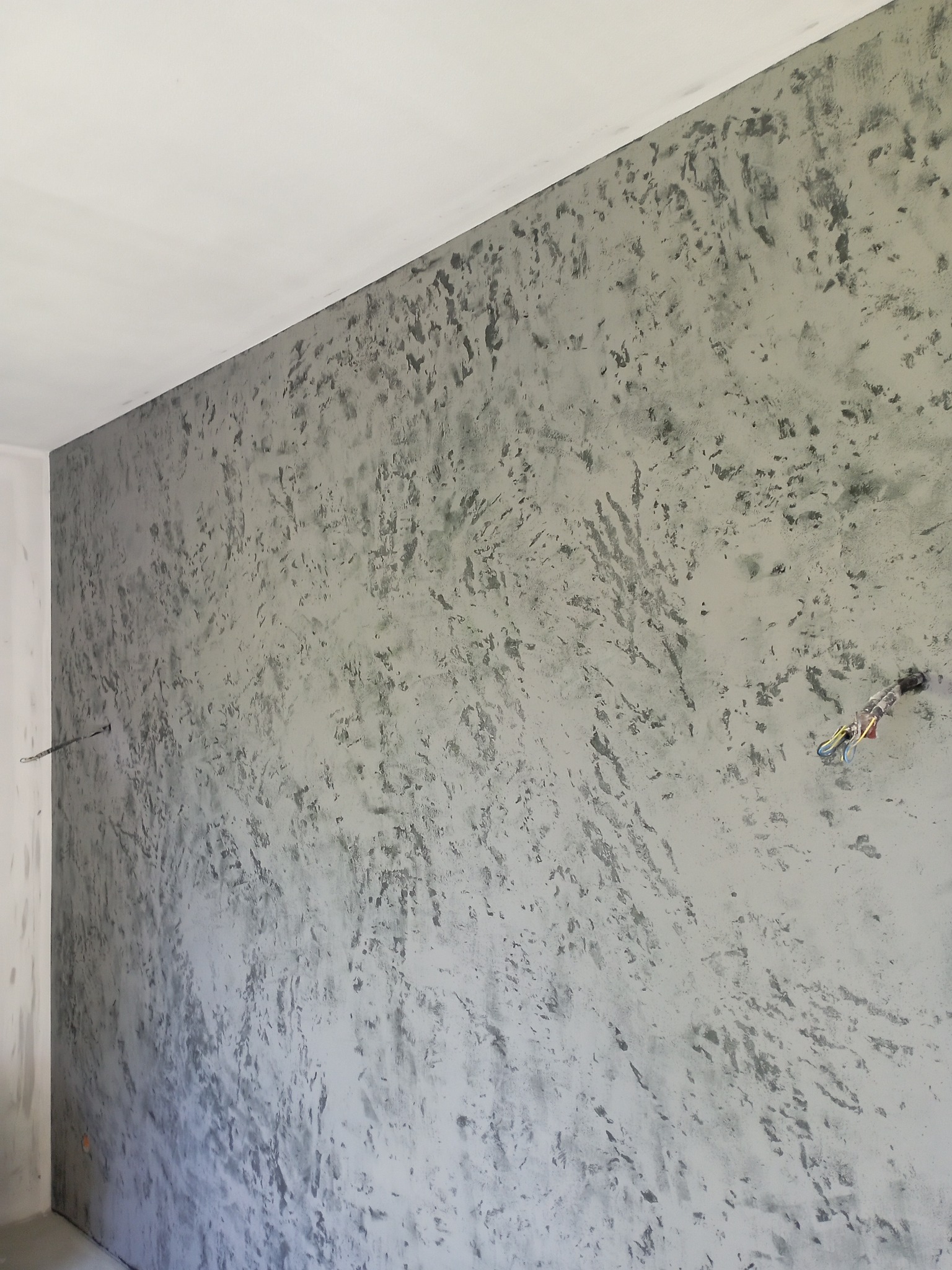 Ściana z dekoracyjnym, szarym tynkiem strukturalnym o nieregularnym wzorze. Widoczne kable elektryczne wystające ze ściany. Biały sufit w rogu pomieszczenia.