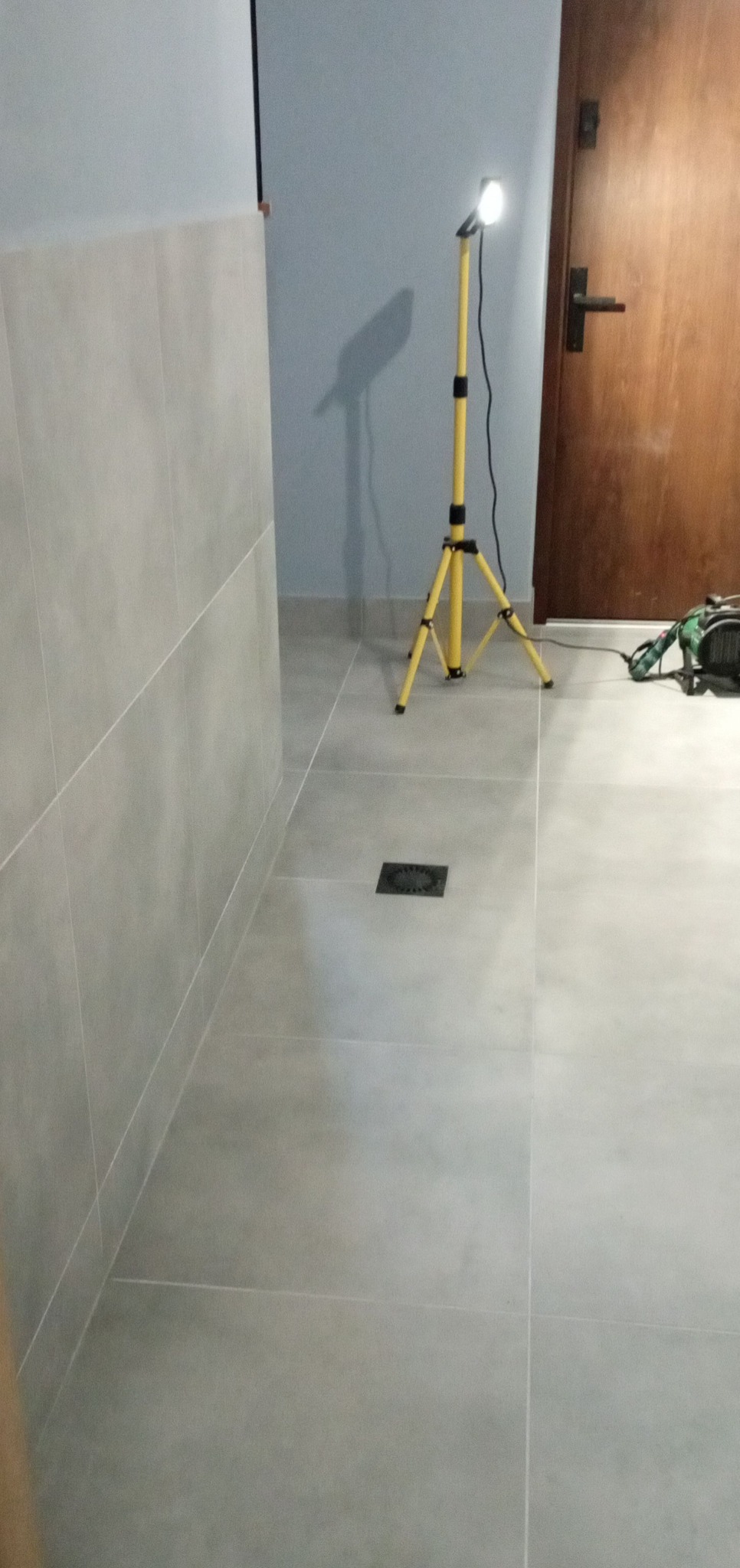 Wykończona łazienka z szarymi płytkami, oświetlona lampą na statywie, z widocznym odpływem liniowym. Widoczny fragment drzwi i ściany.