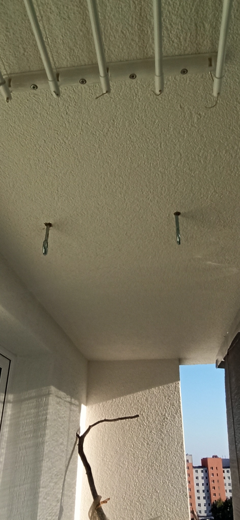 Biały sufit balkonu z zamontowaną suszarką i haczykami, ściany w jasnym odcieniu, widok na budynek w tle.