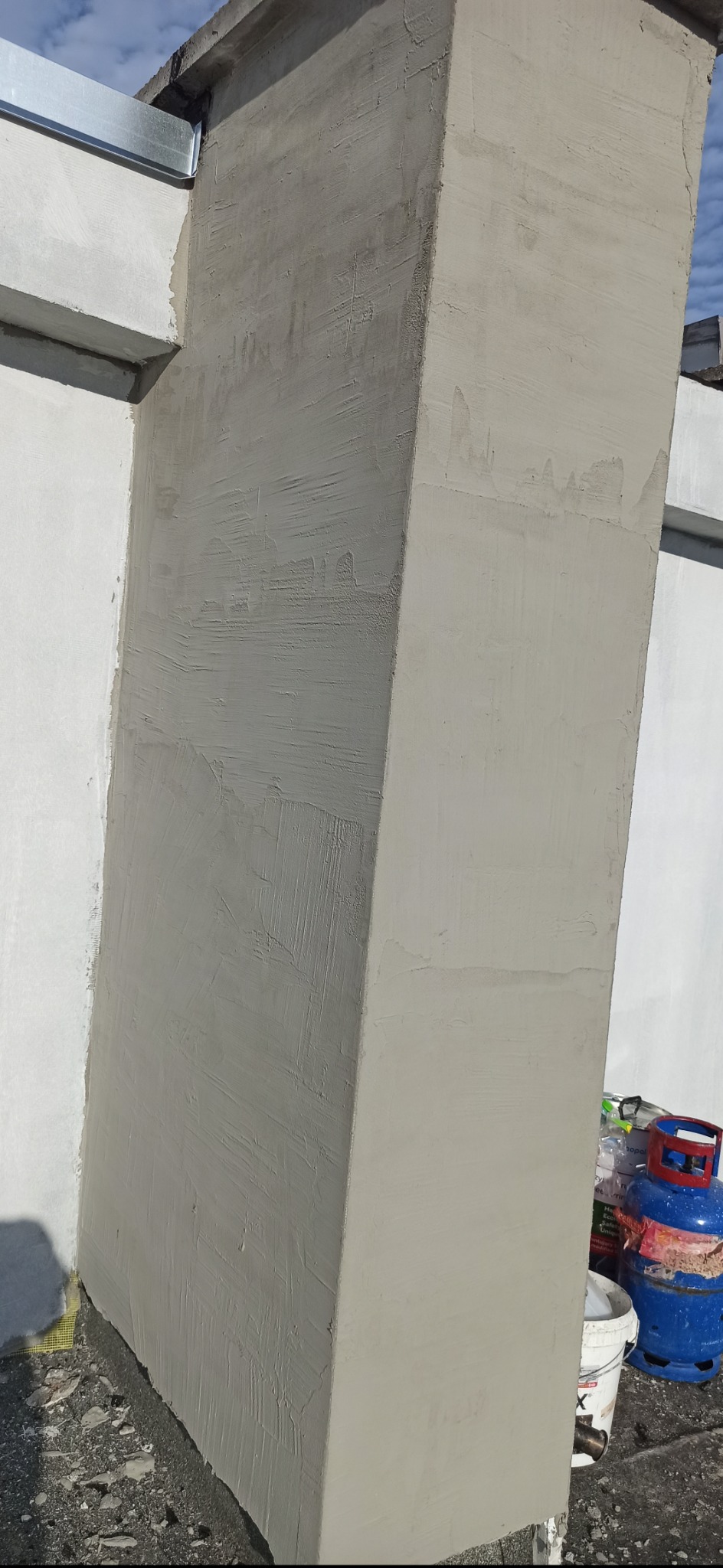 Świeżo pomalowany narożnik budynku z widoczną fakturą farby, obok butla gazowa i wiaderko. Prace wykończeniowe elewacji w trakcie realizacji.