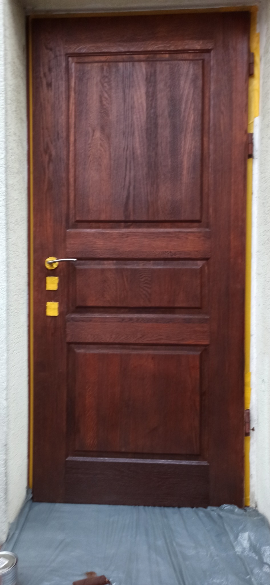 Odnowione, brązowe drzwi z maskowaniem na framudze i folią ochronną na podłodze. Widoczny uchwyt i puszka z farbą w dolnym rogu. Detal po renowacji.