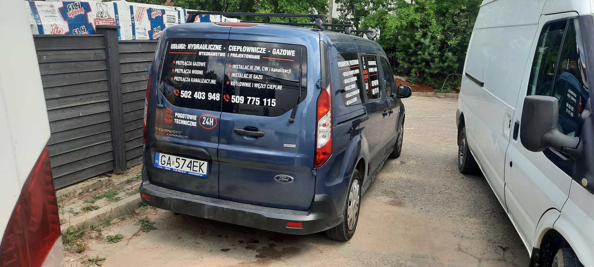 Niebieski Ford Transit Connect z reklamą usług hydraulicznych, gazowych i ciepłowniczych na tylnej szybie. Widoczne numery kontaktowe i logo pogotowia technicznego.