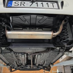 MarioExpert - Podwozie samochodu po konserwacji antykorozyjnej, widoczne sprężyny, wydech i elementy zawieszenia pokryte czarnym preparatem, auto na podnośniku.