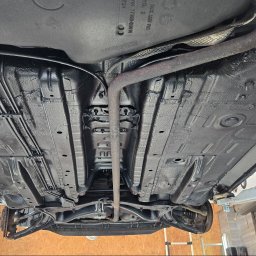 MarioExpert - Podwozie Opel po konserwacji antykorozyjnej, widoczne z dołu na podnośniku. Całość pokryta czarną, ochronną powłoką, z widocznymi elementami układu wydechowego i przewodów.