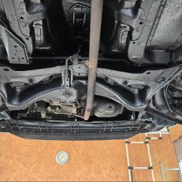 MarioExpert - Podwozie samochodu po zabezpieczeniu antykorozyjnym, widoczne elementy zawieszenia i układu wydechowego. Świeża, czarna powłoka ochronna na metalowych elementach.