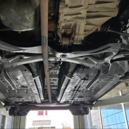 MarioExpert - Podwozie auta po konserwacji antykorozyjnej, widok od dołu na podnośniku. Elementy zawieszenia i podwozia pokryte czarną powłoką ochronną.