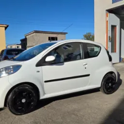 Biały samochód po lakierowaniu z czarnymi felgami w Bujakowie, stojący przed lakiernią. Widoczne budynki w tle i fragment drugiego auta.