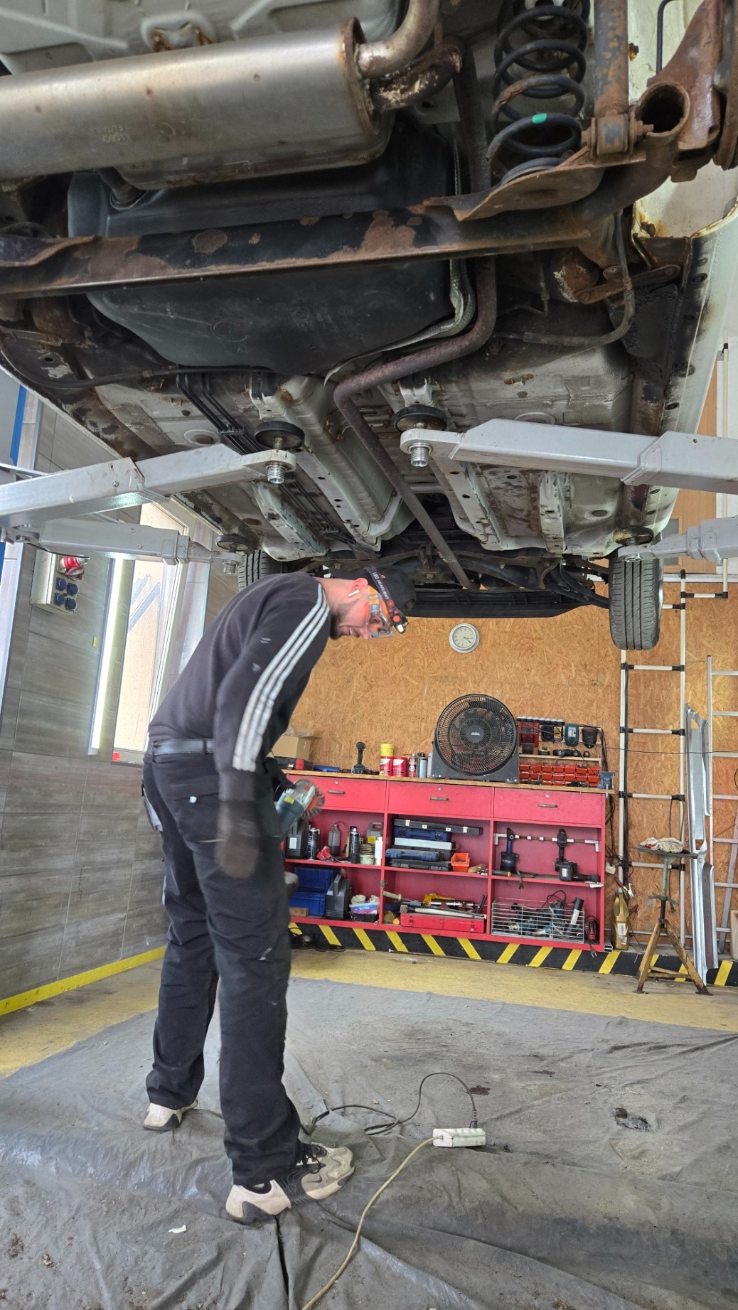 Mechanik w warsztacie, podnosi samochód na podnośniku, przygotowuje się do pracy z użyciem narzędzia elektrycznego. Widoczne elementy podwozia pojazdu.