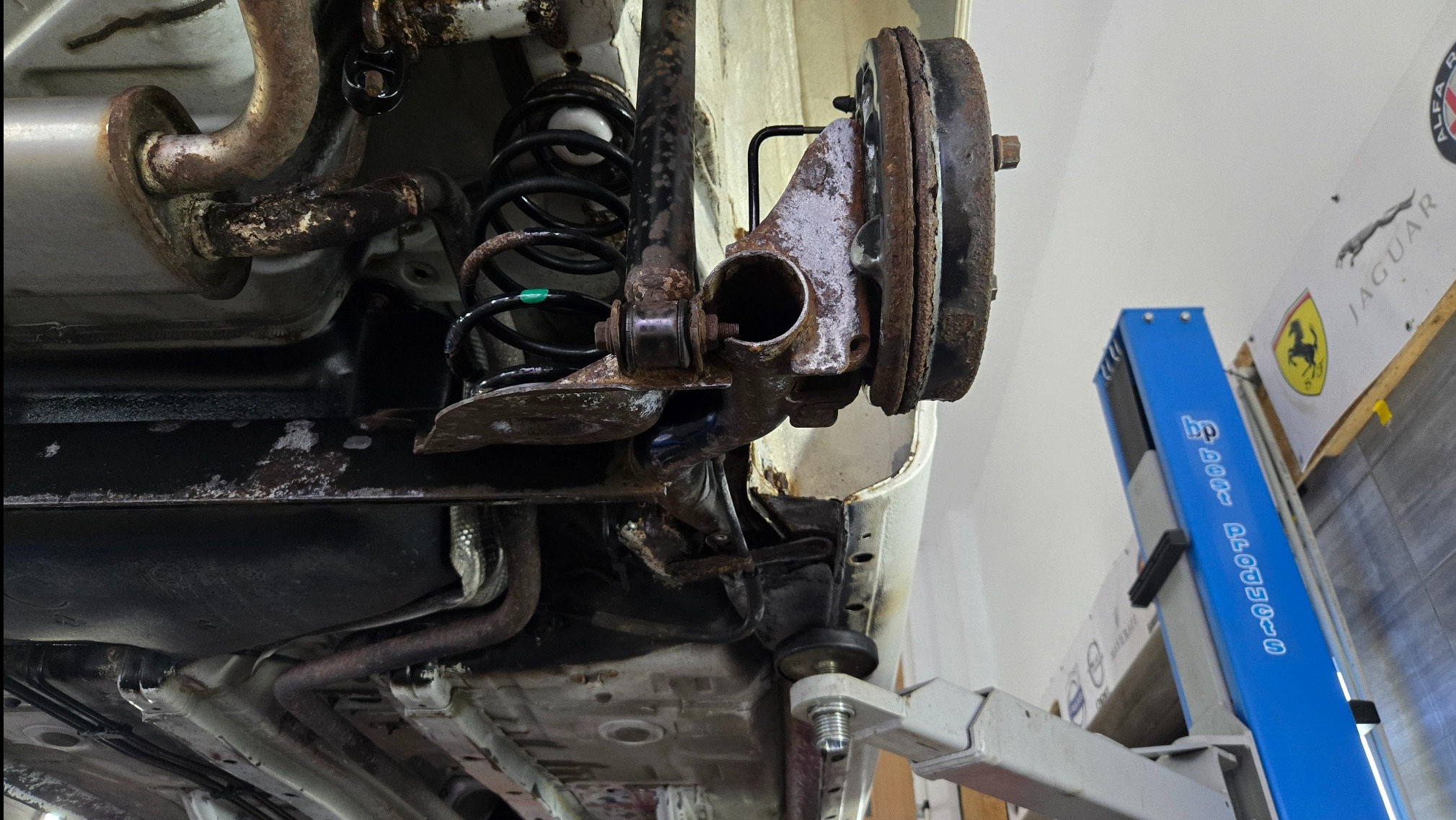 Podwozie skorodowanego samochodu na podnośniku warsztatowym, widoczne elementy zawieszenia, sprężyna i układ wydechowy. W tle logo marek Jaguar i Ferrari.