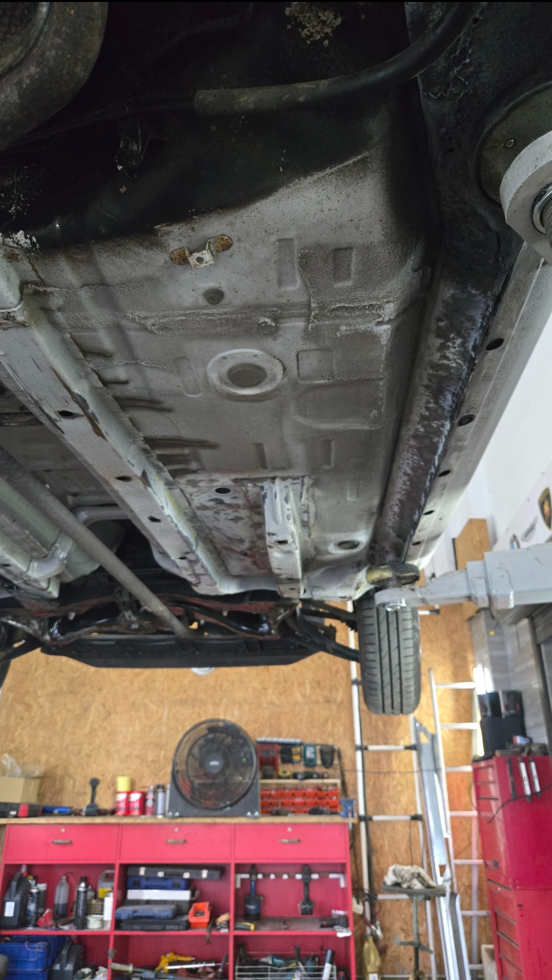 Podwozie samochodu na podnośniku w warsztacie, widoczne elementy zawieszenia i skorodowane fragmenty. W tle narzędzia i wyposażenie warsztatowe.