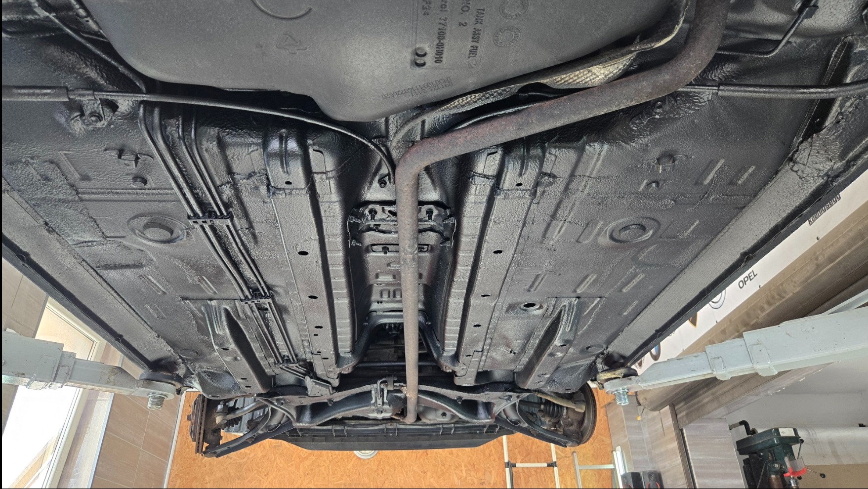 Podwozie Opel po konserwacji antykorozyjnej, widoczne z dołu na podnośniku. Całość pokryta czarną, ochronną powłoką, z widocznymi elementami układu wydechowego i przewodów.