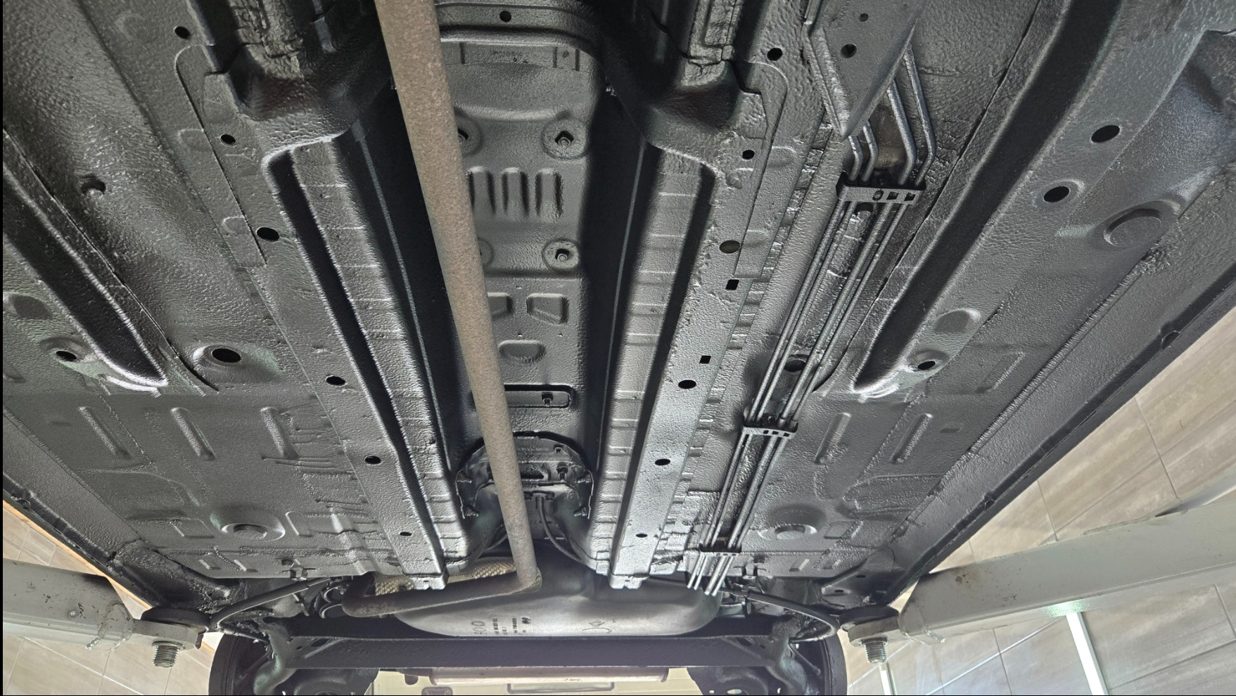 Podwozie samochodu po konserwacji antykorozyjnej, widok od dołu na podnośniku. Elementy pokryte jednolitą, czarną powłoką ochronną. Widoczne przewody i elementy zawieszenia.