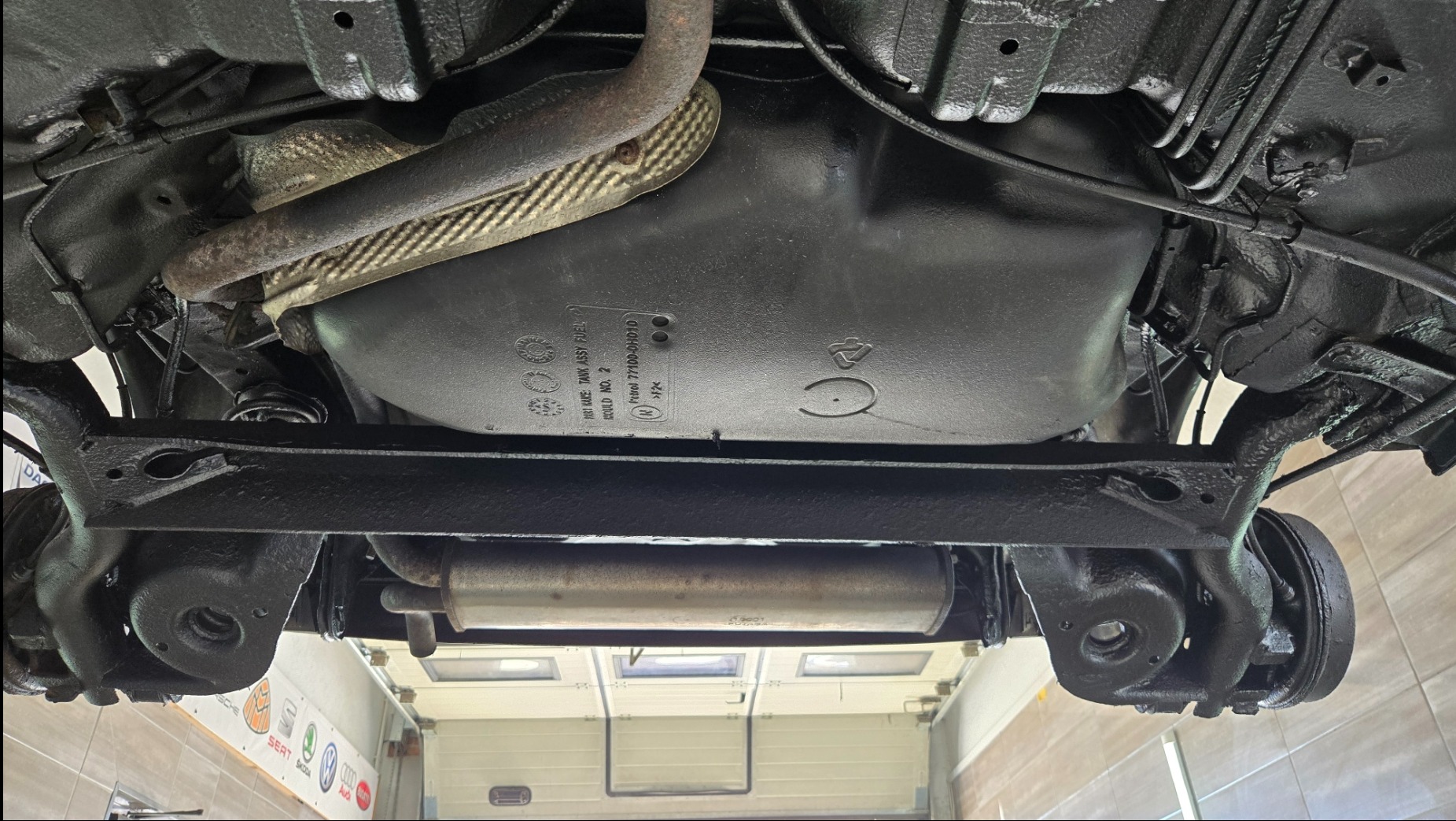 Podwozie auta po konserwacji antykorozyjnej, widok od spodu. Elementy zawieszenia i układu wydechowego pokryte czarną, ochronną powłoką. Widoczny zbiornik paliwa z oznaczeniami.