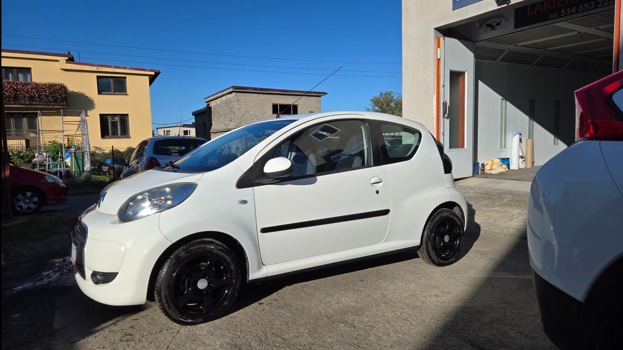 Biały samochód po lakierowaniu z czarnymi felgami w Bujakowie, stojący przed lakiernią. Widoczne budynki w tle i fragment drugiego auta.